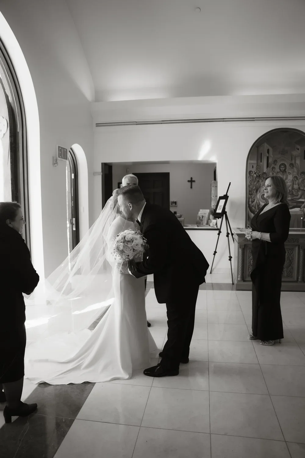 A bride and groom share a kiss inside a church during their wedding ceremony, with a woman standing nearby and a officiant in the background.