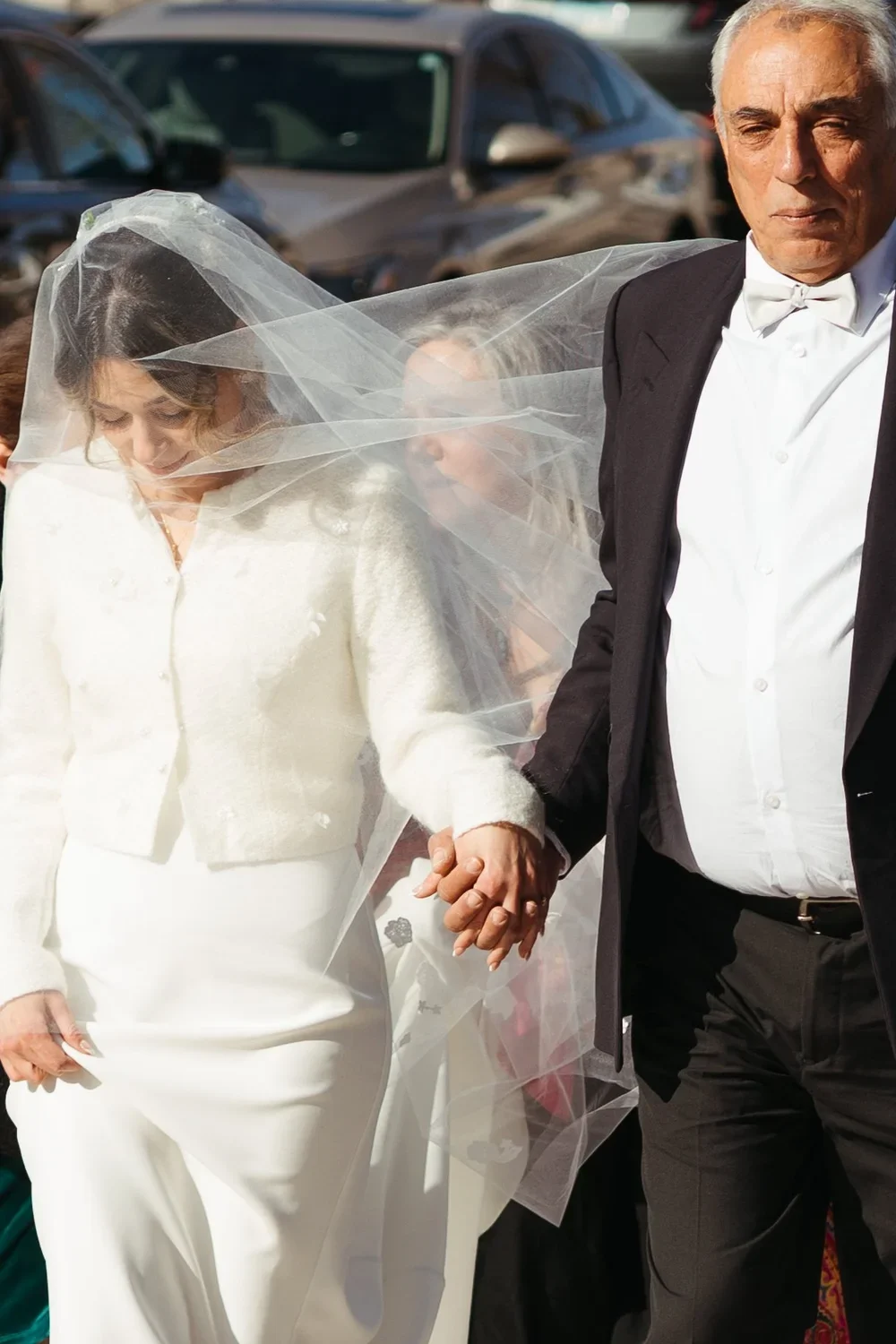 A bride and groom holding hands at their wedding ceremony outdoors, with a blurred parking lot in the background. The bride is wearing a white dress and a veil, and the groom is dressed in a tuxedo.