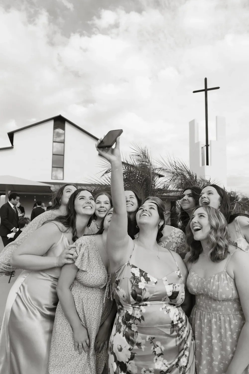 A group of women in formal attire taking a selfie in front of a church with a cross, palm trees, and a partly cloudy sky.