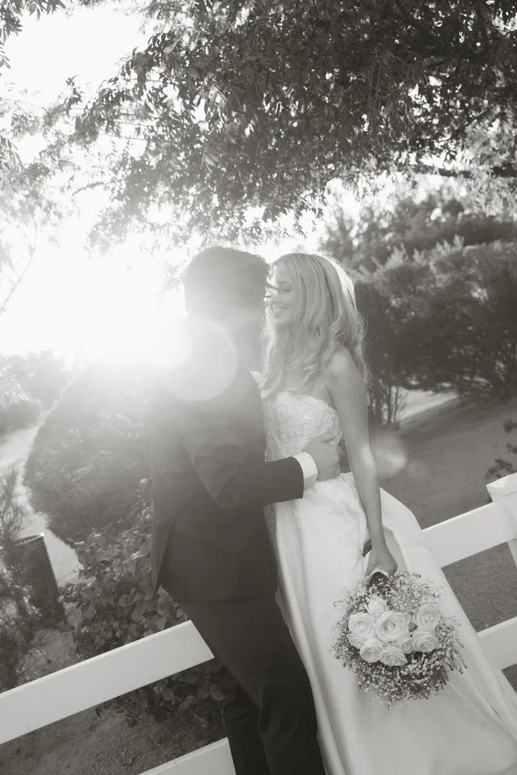 A bride and groom share a kiss outdoors, with sunlight filtering through trees, the bride holding a bouquet of roses.