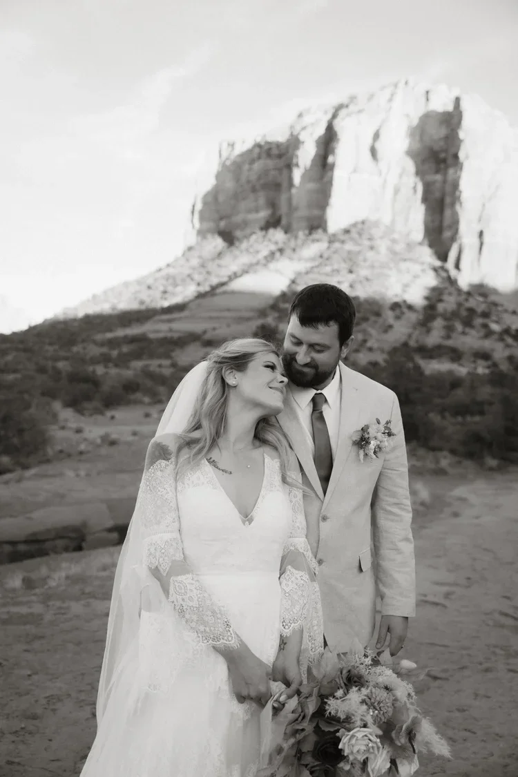 Black and white photo of a bride and groom standing close together outdoors, with a rocky mountain in the background. The bride is holding a bouquet, and they are smiling affectionately at each other.