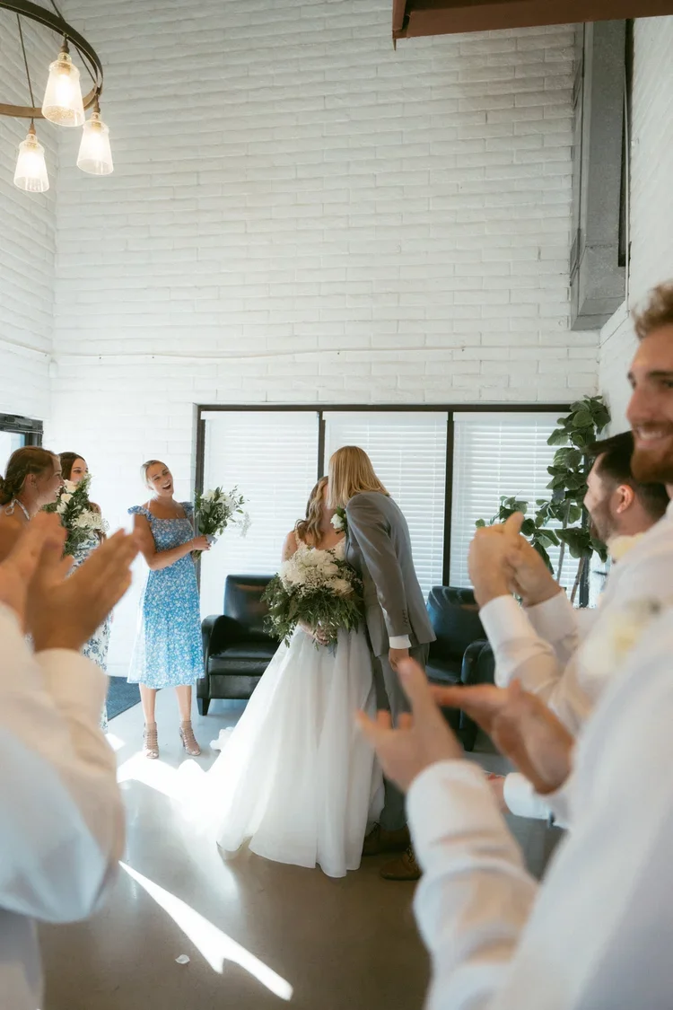 A bride and groom sharing a kiss during their wedding ceremony with friends and family clapping and celebrating in a bright, modern room.