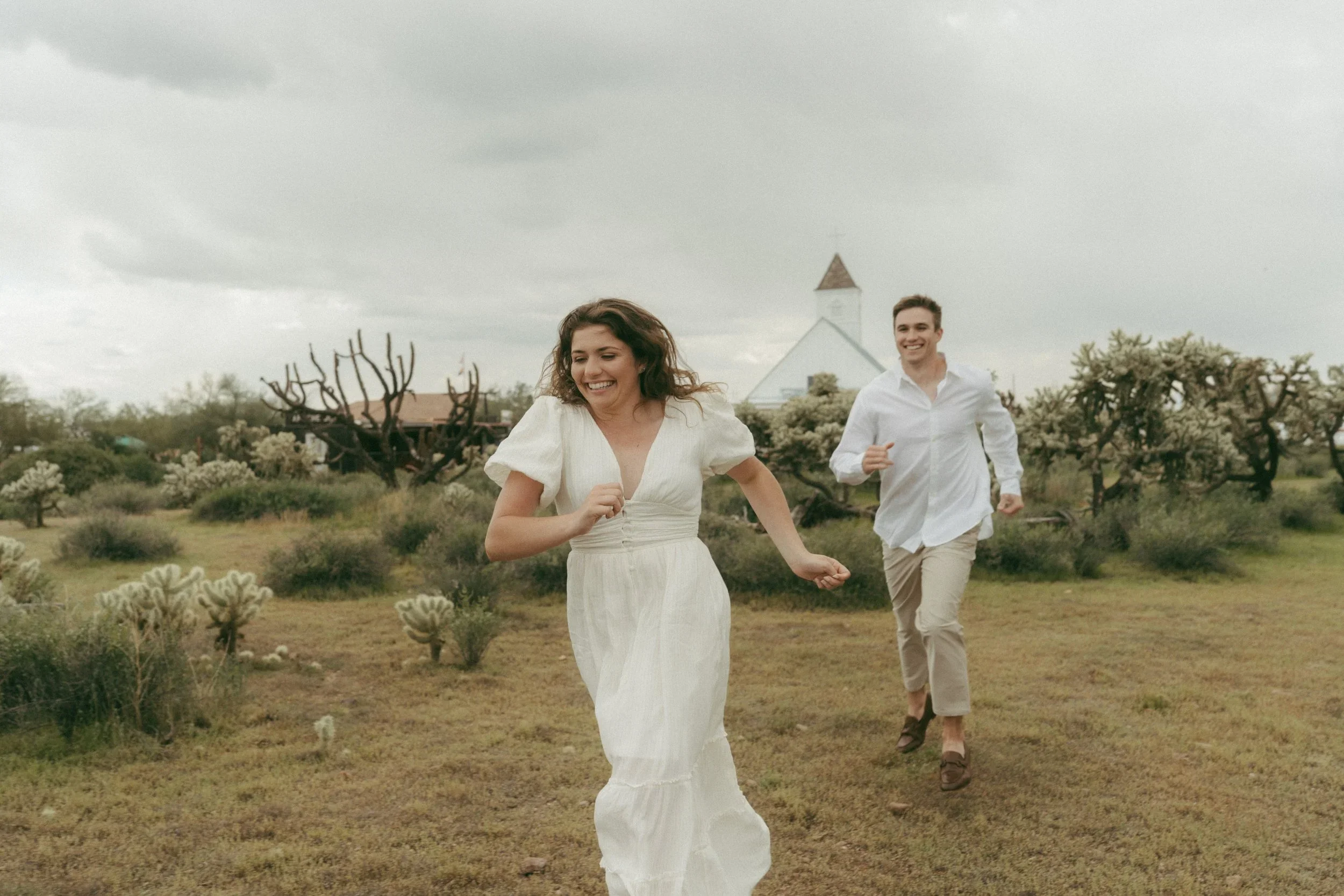 A woman and a man running outdoors in a field with various trees and bushes under a cloudy sky. The woman is smiling and wearing a white dress, while the man is also smiling and dressed in a white shirt and beige pants.