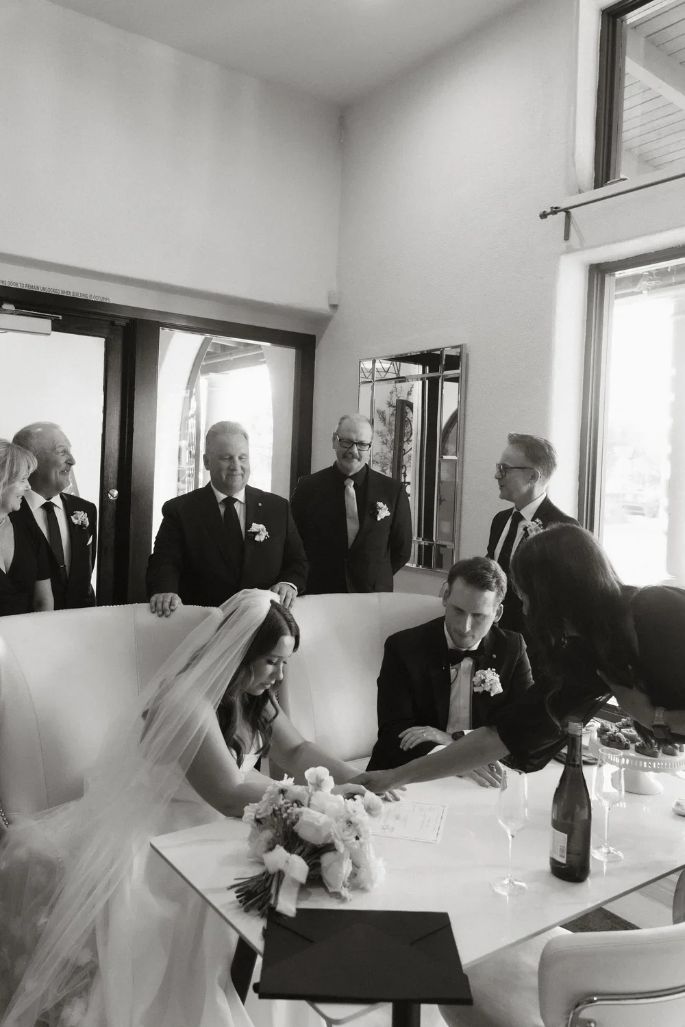 A black-and-white photo of a wedding ceremony, with a bride and groom sitting at a table, signing documents. The bride is wearing a veil and flower bouquet, and the groom is in a tuxedo. Several people, dressed in formal attire, are standing and watching in the background inside a well-lit room with large windows.