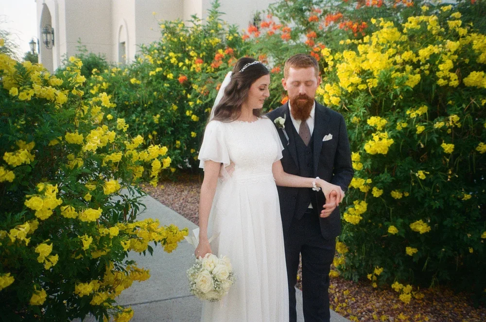 A bride and groom walking together on a pathway, surrounded by vibrant yellow and orange flowers, during a wedding photoshoot.