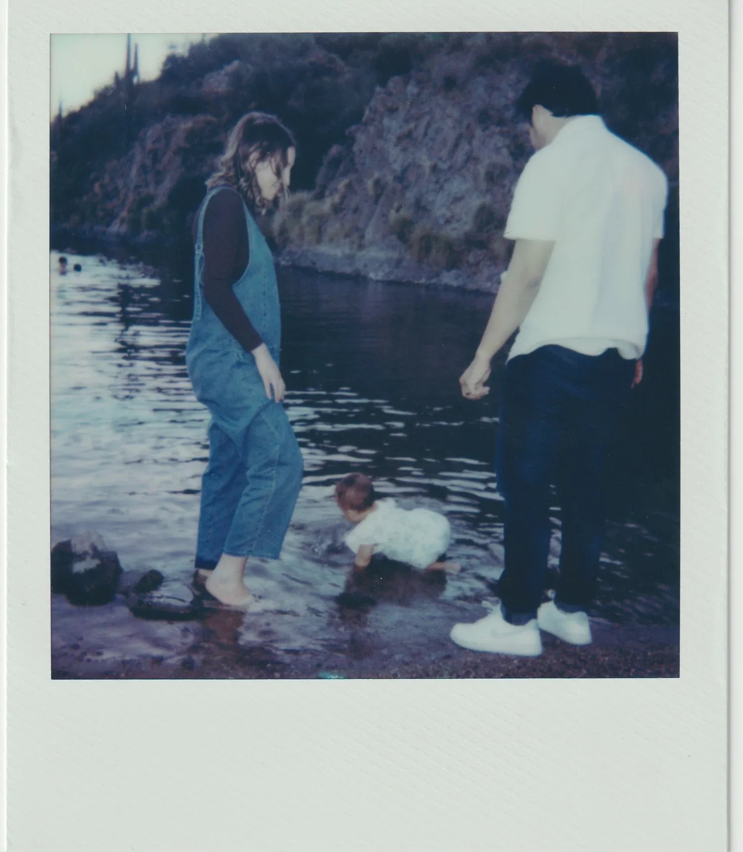 Four people wading in a river near rocky cliffs, two standing and leaning over, one child's head visible in the water, and another person sitting on the shore.