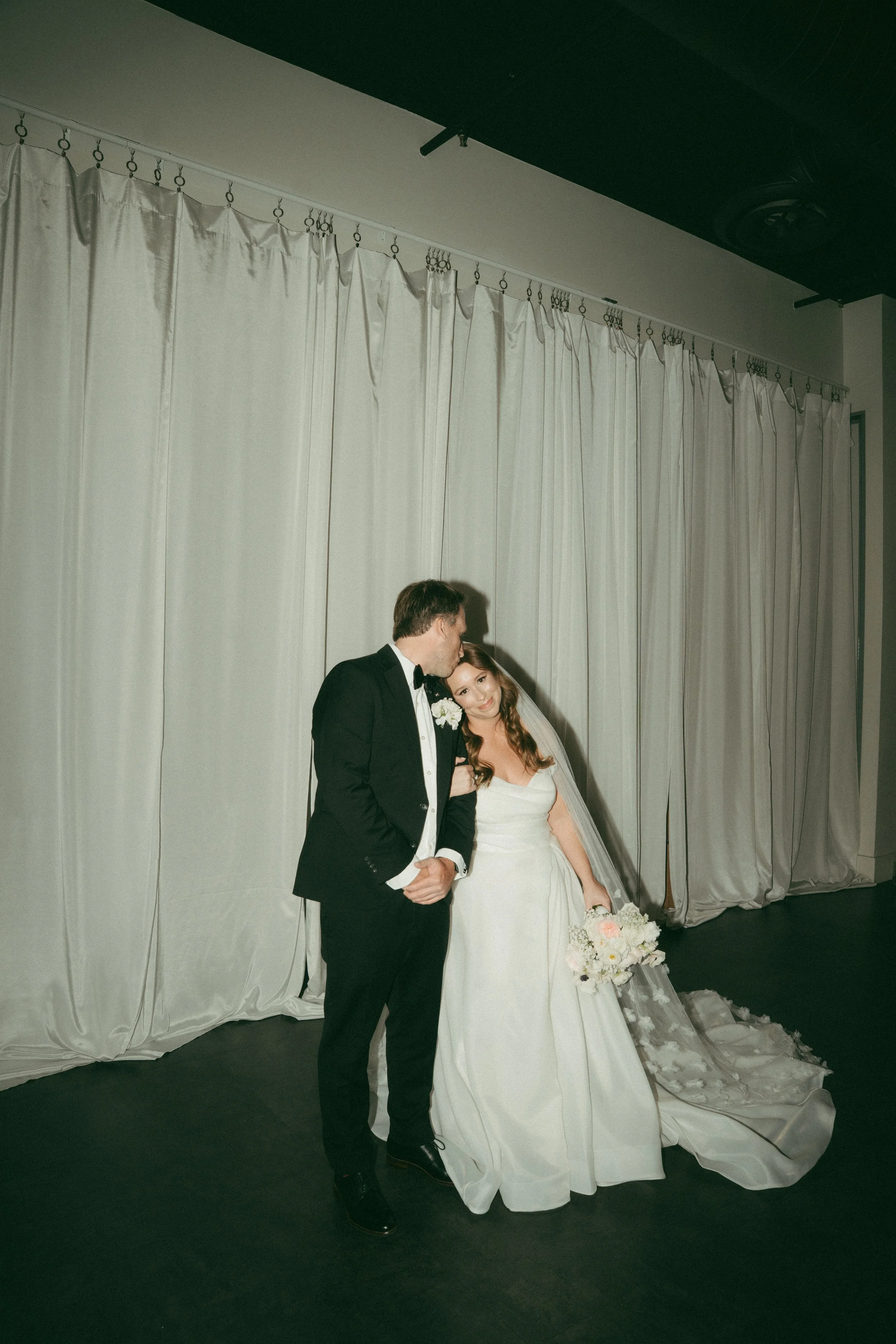 A bride and groom standing together, with the groom kissing the bride's forehead. The bride is holding a bouquet of flowers and is wearing a white wedding gown. They are in front of white curtains at a wedding venue.