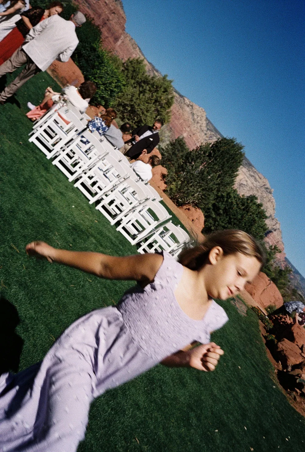 Young girl running or playing outside on a grassy area during an outdoor event with white chairs arranged in a row and people gathering in the background, set against a backdrop of mountains and trees.