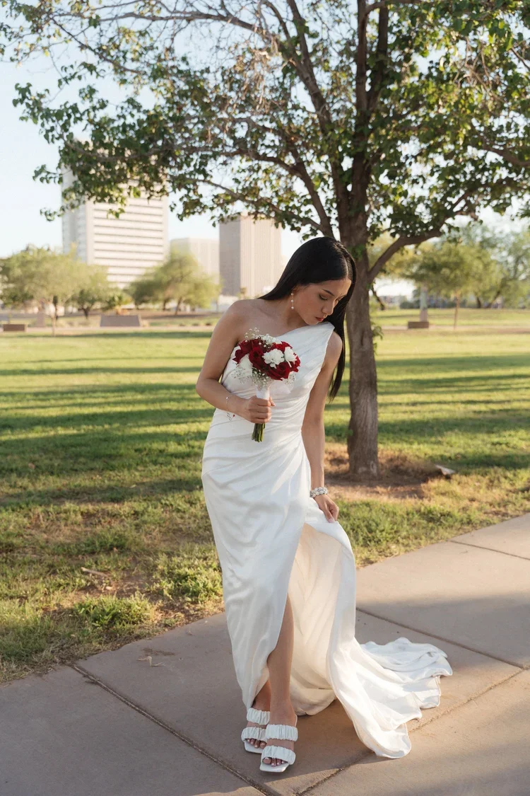 A bride in a white wedding dress holding a bouquet of red and white flowers, standing on a sidewalk near a grassy park with trees and buildings in the background.