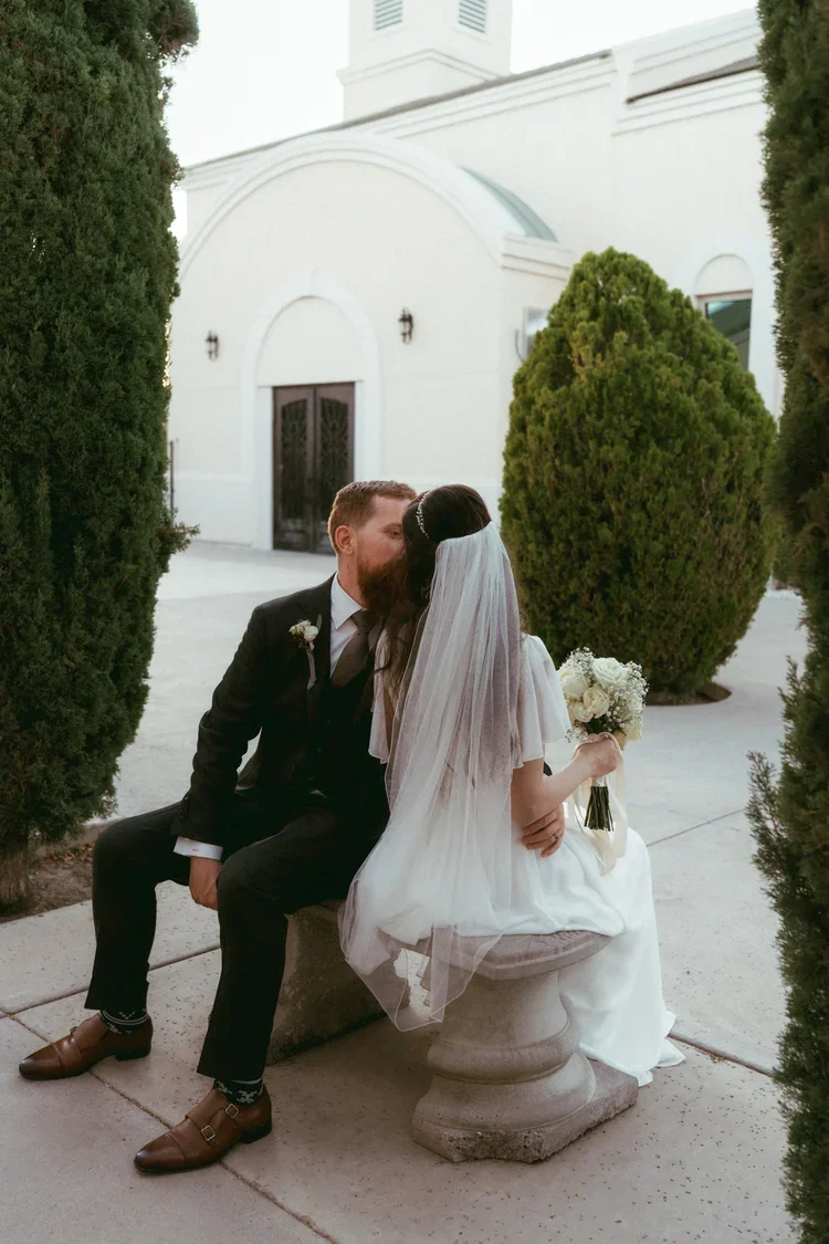 A bride and groom sitting on a stone bench outside a white building, sharing a kiss. The bride is in a white wedding dress with a veil, holding a bouquet of white roses. The groom is in a black suit with brown shoes.