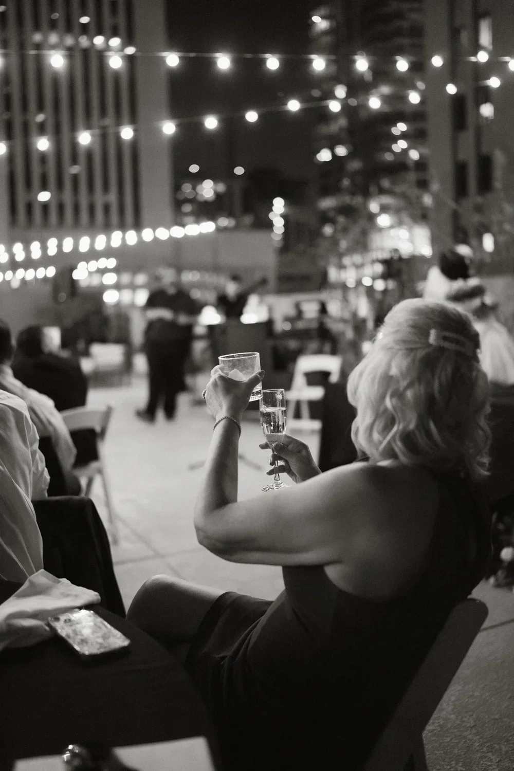 A woman with blonde hair in a black sleeveless dress raising her glass of champagne at an outdoor celebration at night, decorated with string lights and with people and buildings in the background.