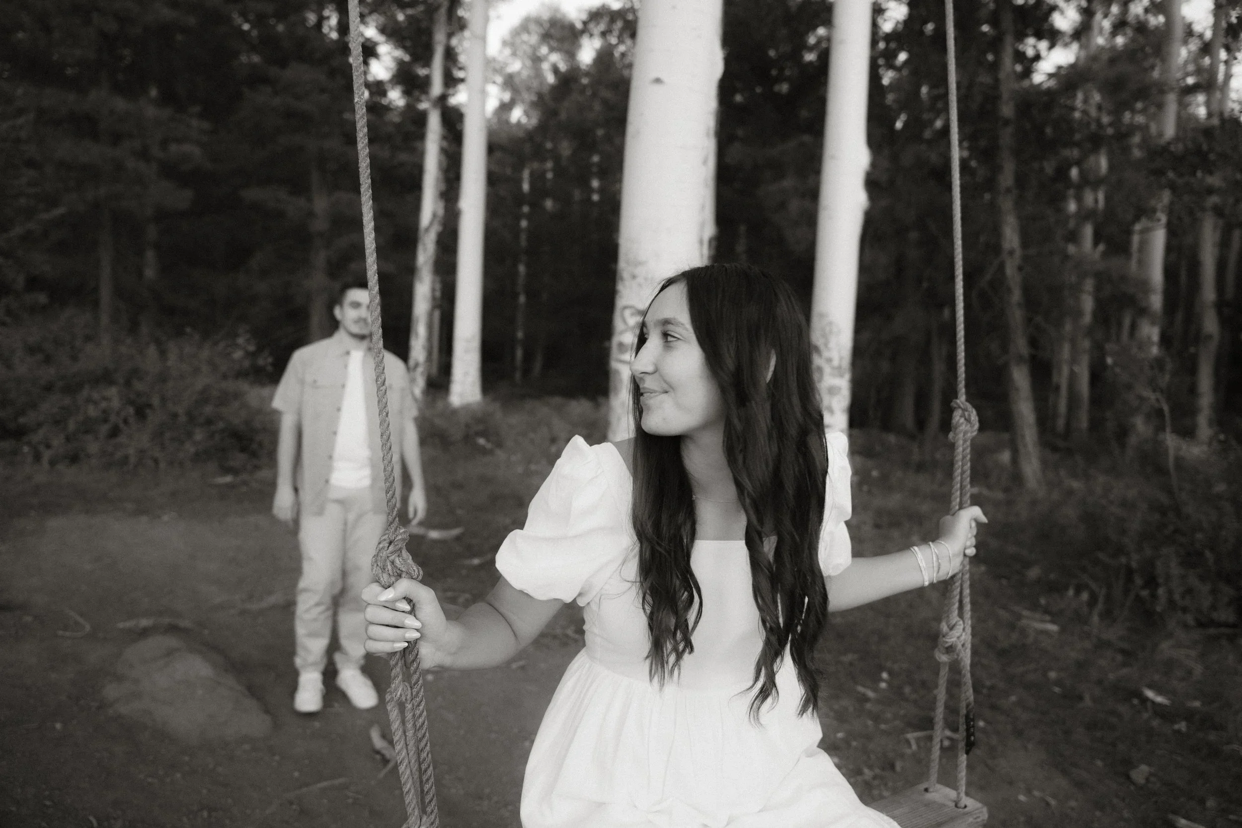 A young woman with long dark hair sitting on a swing and smiling while looking at a young man standing in the background. The scene is outdoors with trees around.