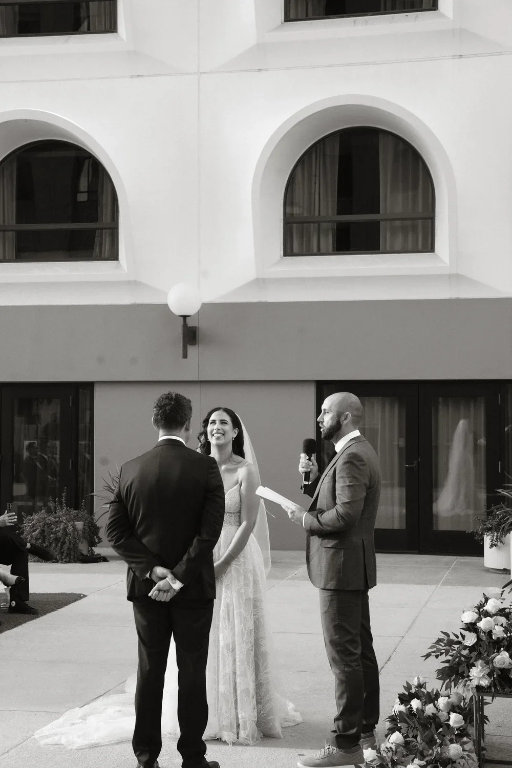 A black and white photo of a wedding ceremony taking place outdoors, with a bride and groom facing each other and a officiant speaking into a microphone.