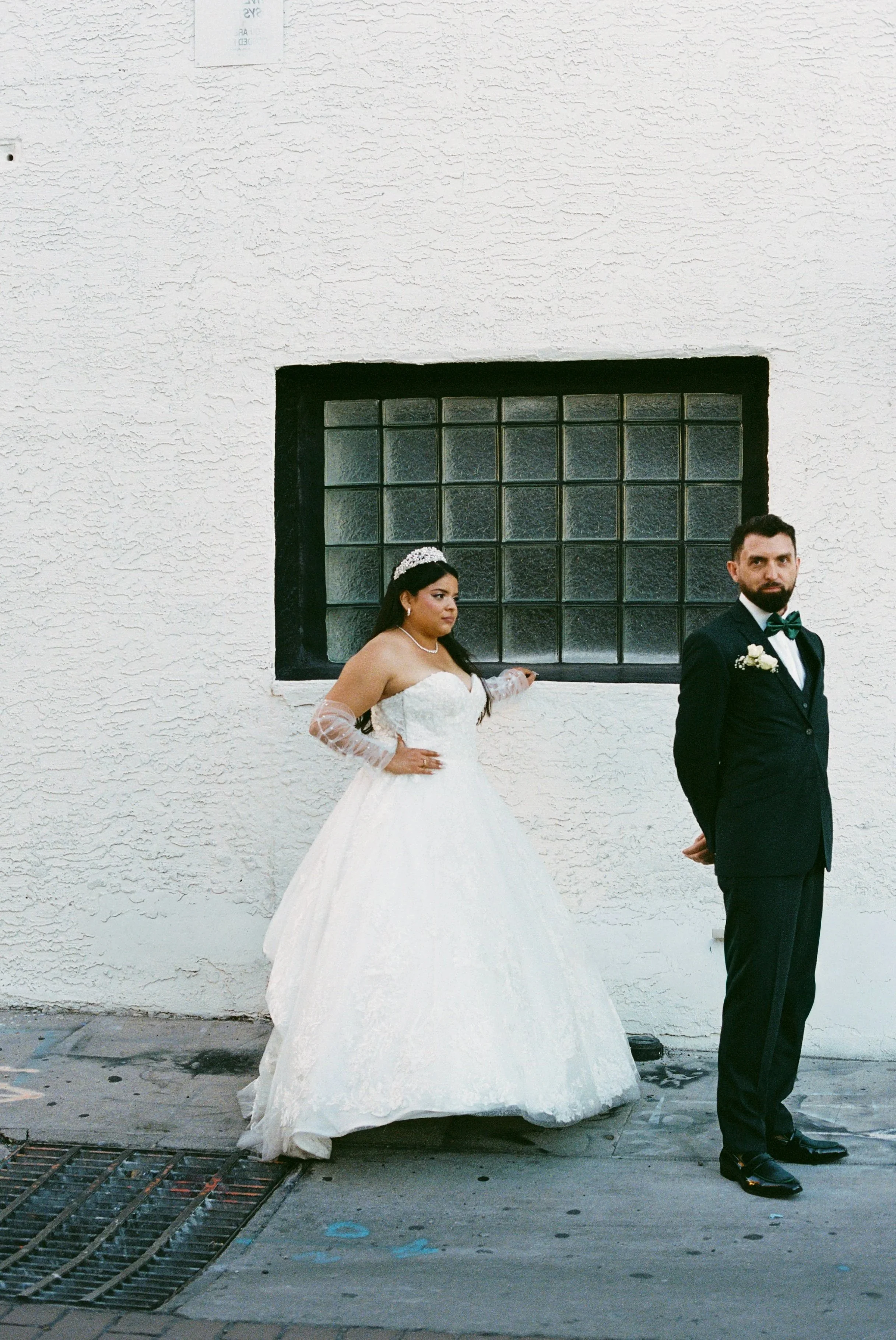 A bride and groom in formal wedding attire standing against a white textured wall with a black-framed glass block window. The bride is wearing a white strapless wedding gown with long lace gloves, a pearl necklace, and a tiara. The groom is dressed in a black tuxedo with a green bow tie and boutonnière, looking to the camera with his hands behind his back.