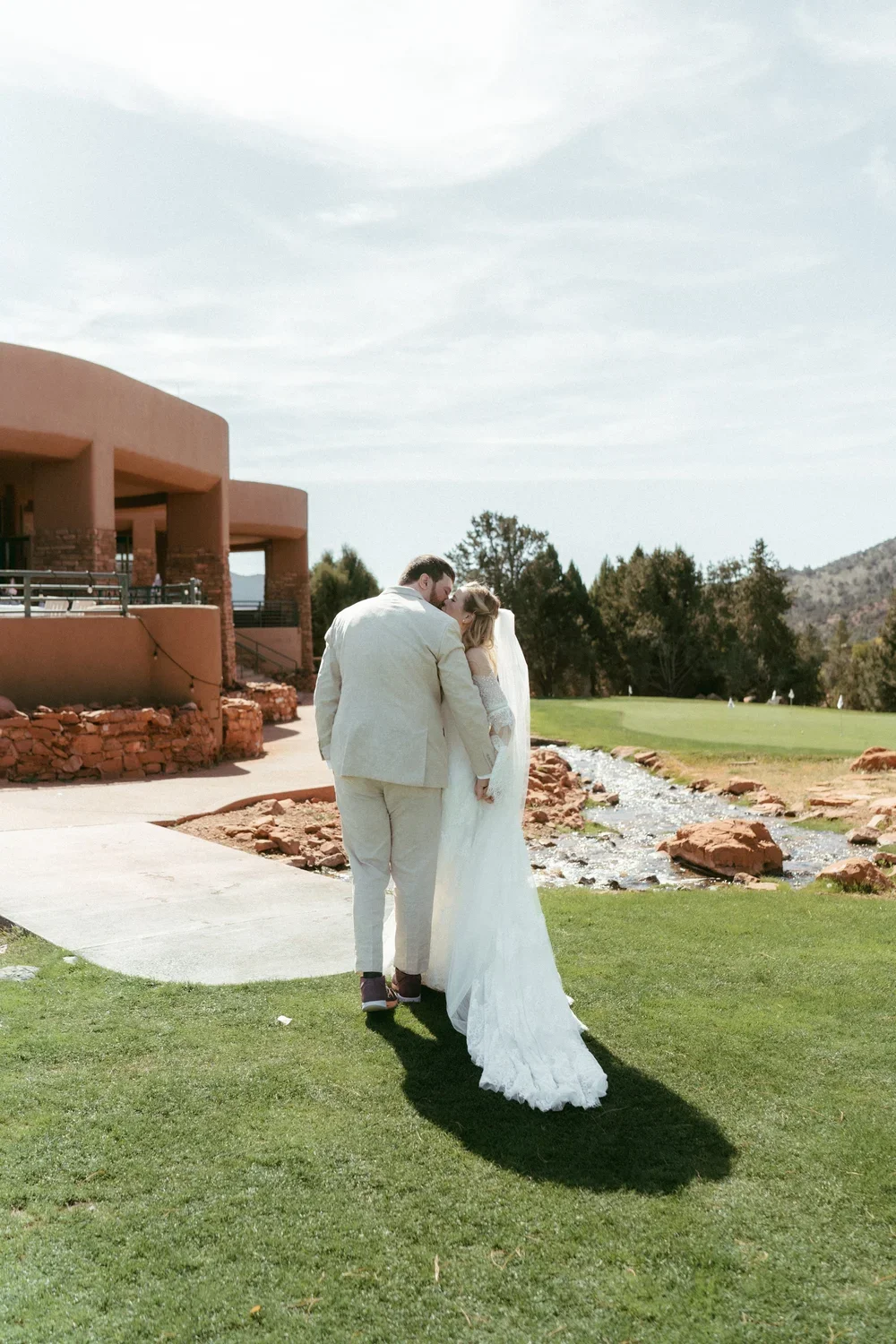 A bride and groom standing on a lawn, sharing a kiss with a scenic natural background including a stream, trees, and distant hills.