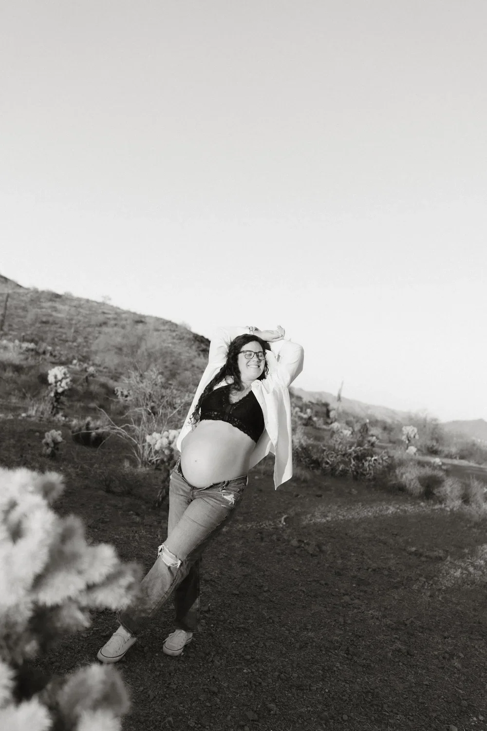 A woman standing outdoors in a desert landscape with cacti, smiling and posing with her hands behind her head, wearing glasses, a black top, an open white shirt, ripped jeans, and sneakers.