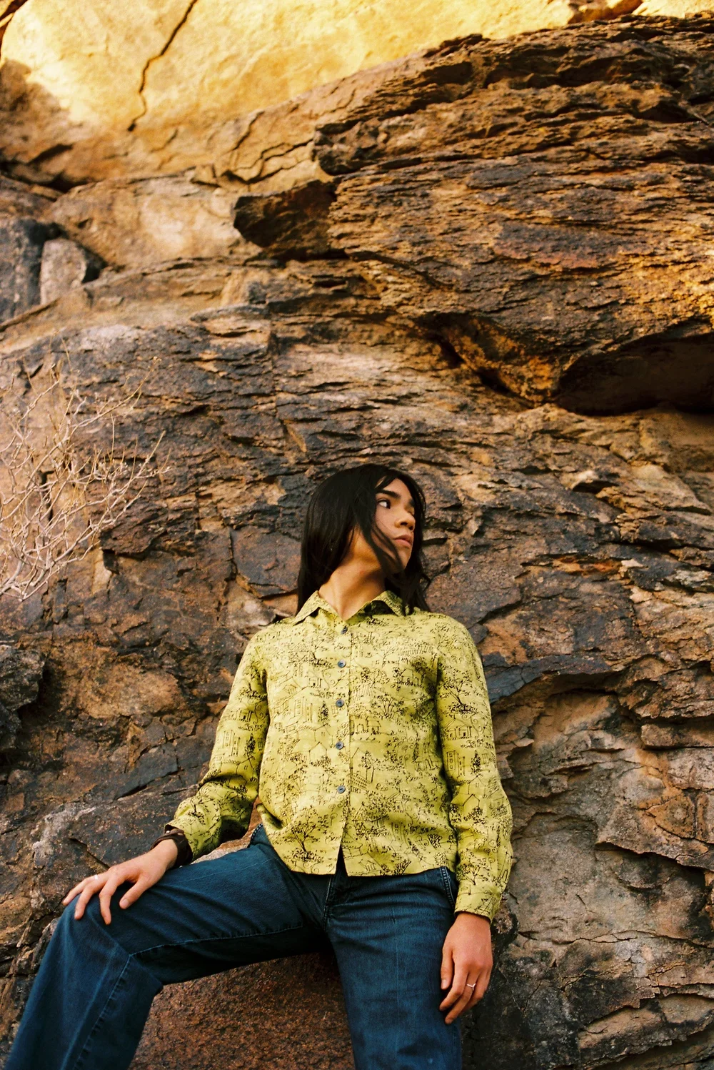 A young woman with long black hair wearing a yellow patterned shirt and blue jeans is standing against a rugged rock wall in a desert landscape.