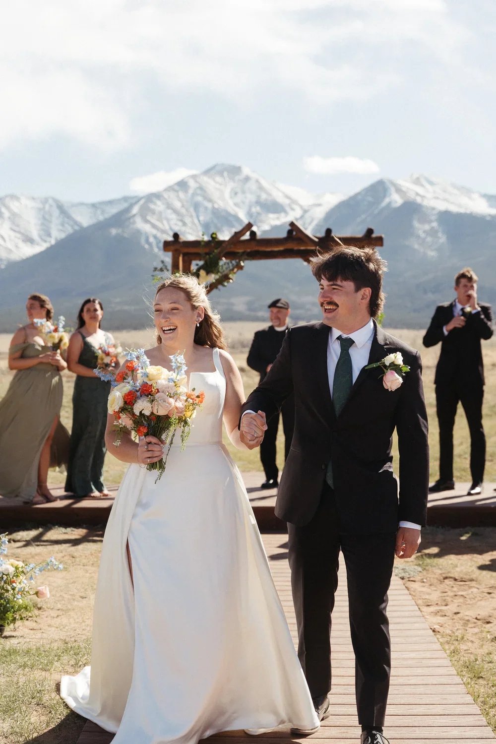 Newlywed couple walking hand-in-hand outdoors, smiling, with a mountain backdrop, wedding guests and a photographer in the background