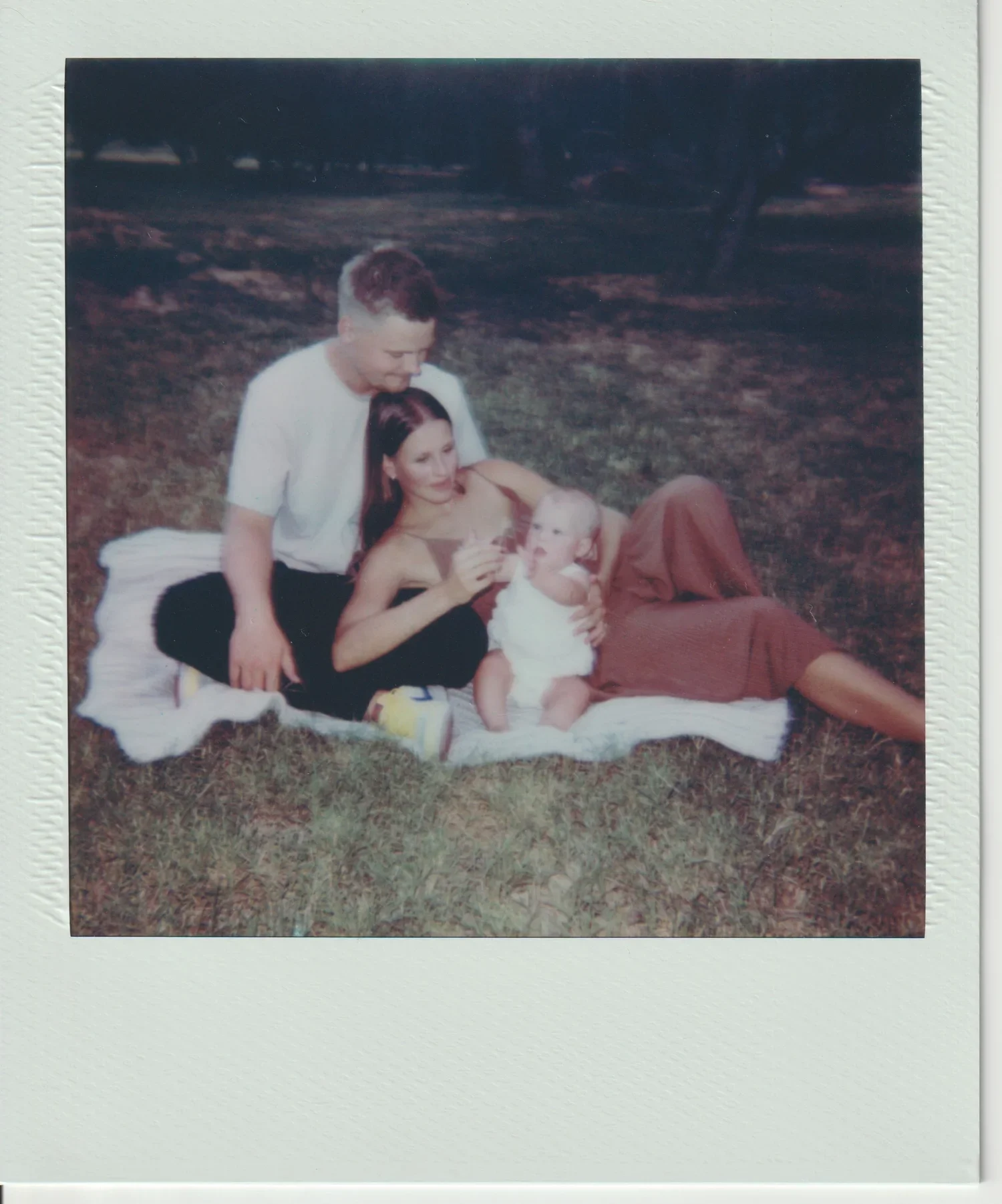A vintage photograph of three people sitting on a blanket outdoors on grass. An adult woman and a young girl are sitting together, while a small child is lying on the blanket. The adult woman is holding the child's hand, and the young girl is looking at the child. It appears to be late afternoon or early evening.