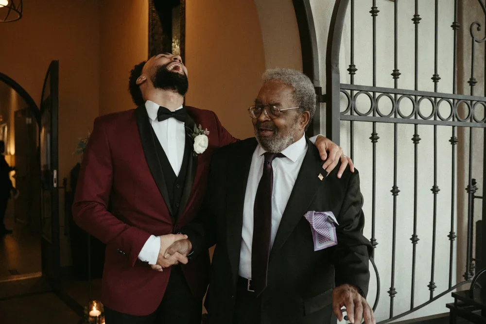 Two men laughing and smiling while shaking hands at a formal event, with one man wearing a burgundy tuxedo and the other in a black suit, in front of a decorative gate.