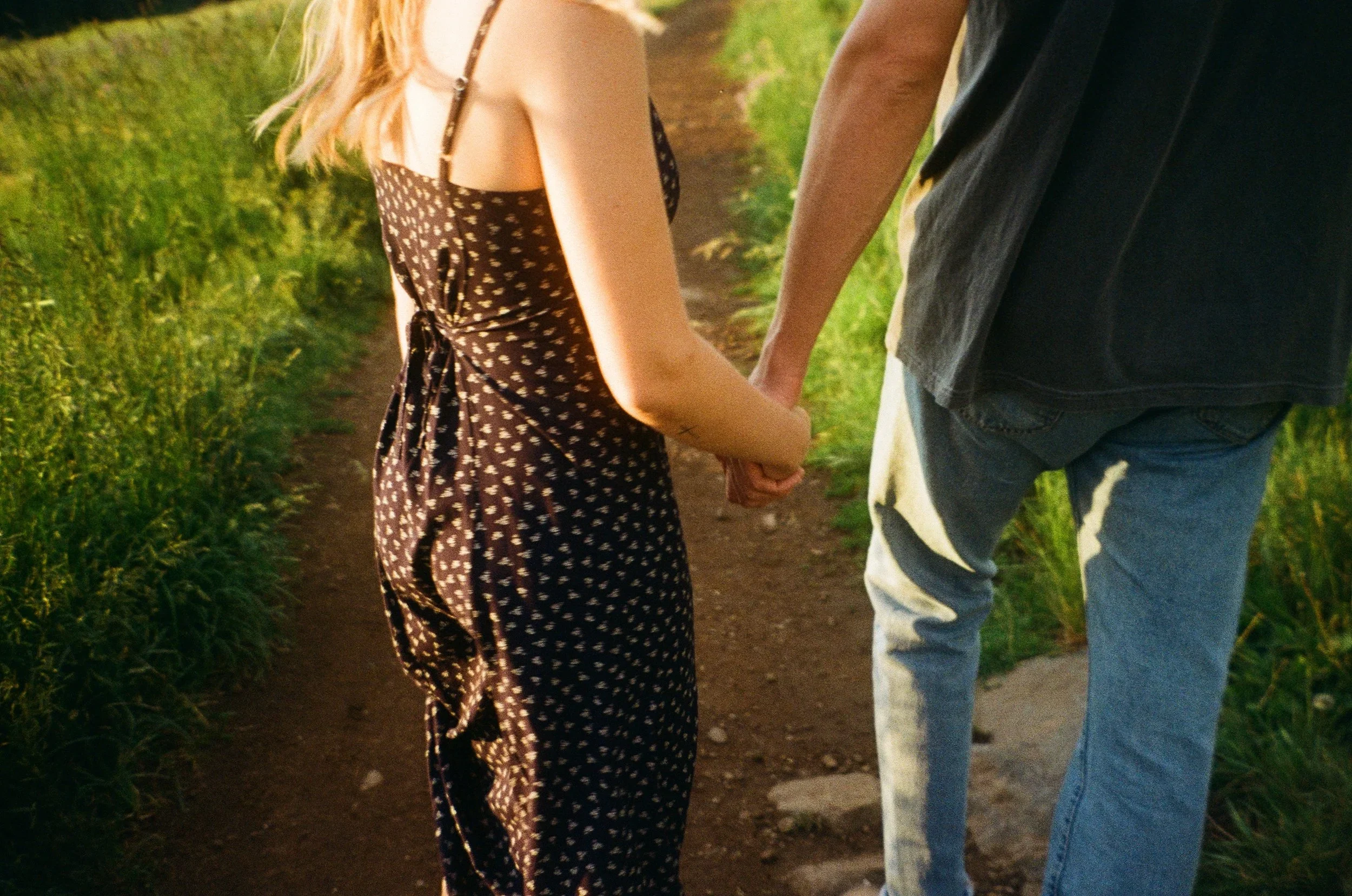 A close-up of a couple holding hands while walking on a dirt path in a green outdoor setting, with the woman's face not visible and the man's face partially visible, during sunset.
