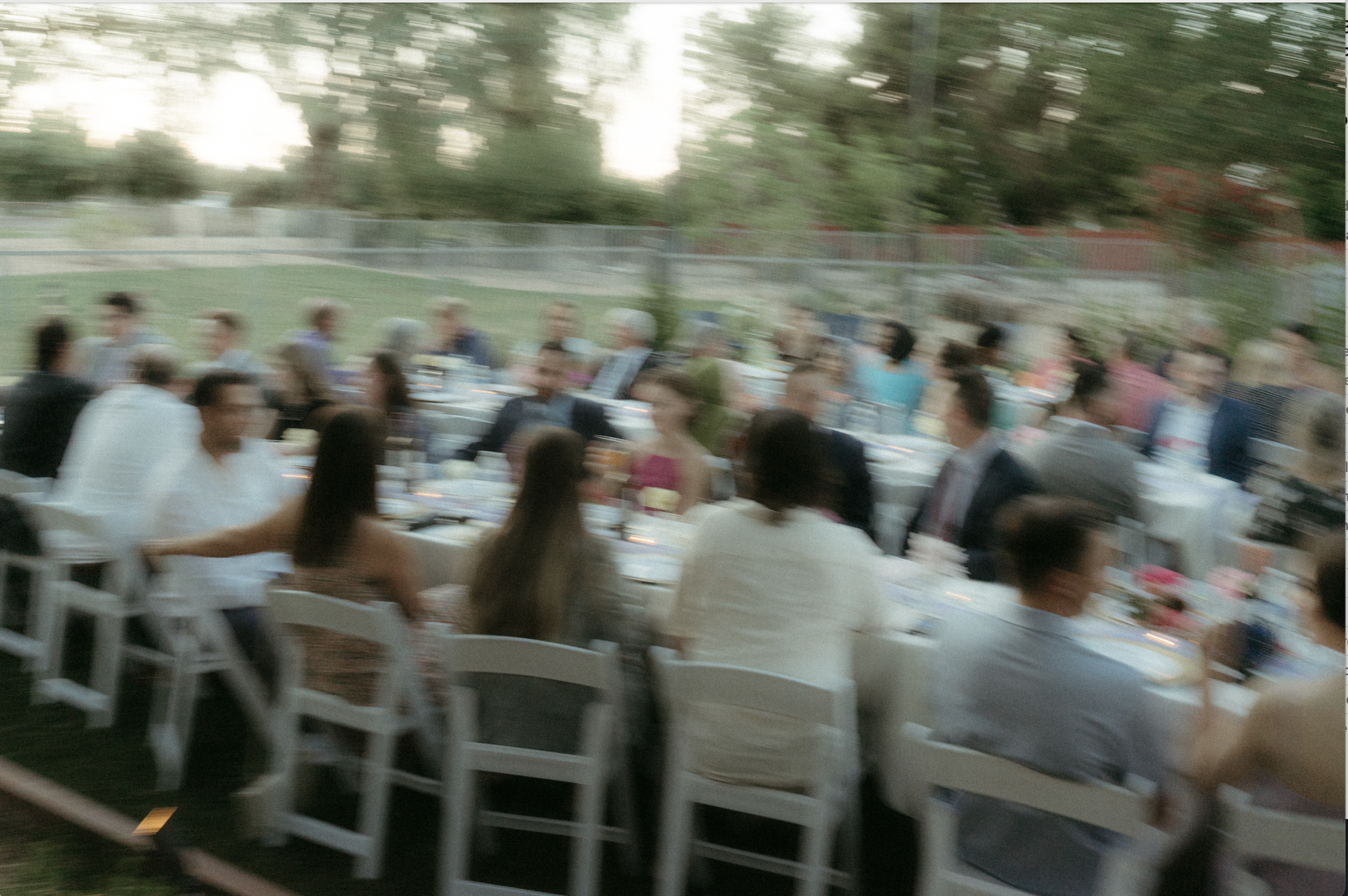 People sitting at long tables outside, with trees and a fence in the background, in a blurred motion effect.