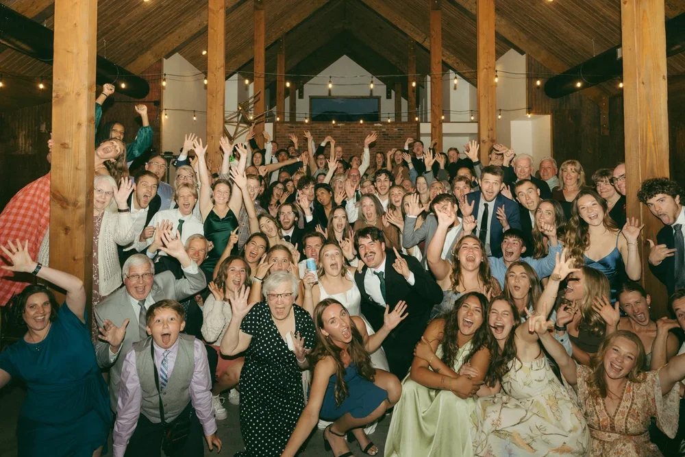 A large group of people gathered inside a wooden barn, celebrating and smiling at the camera.
