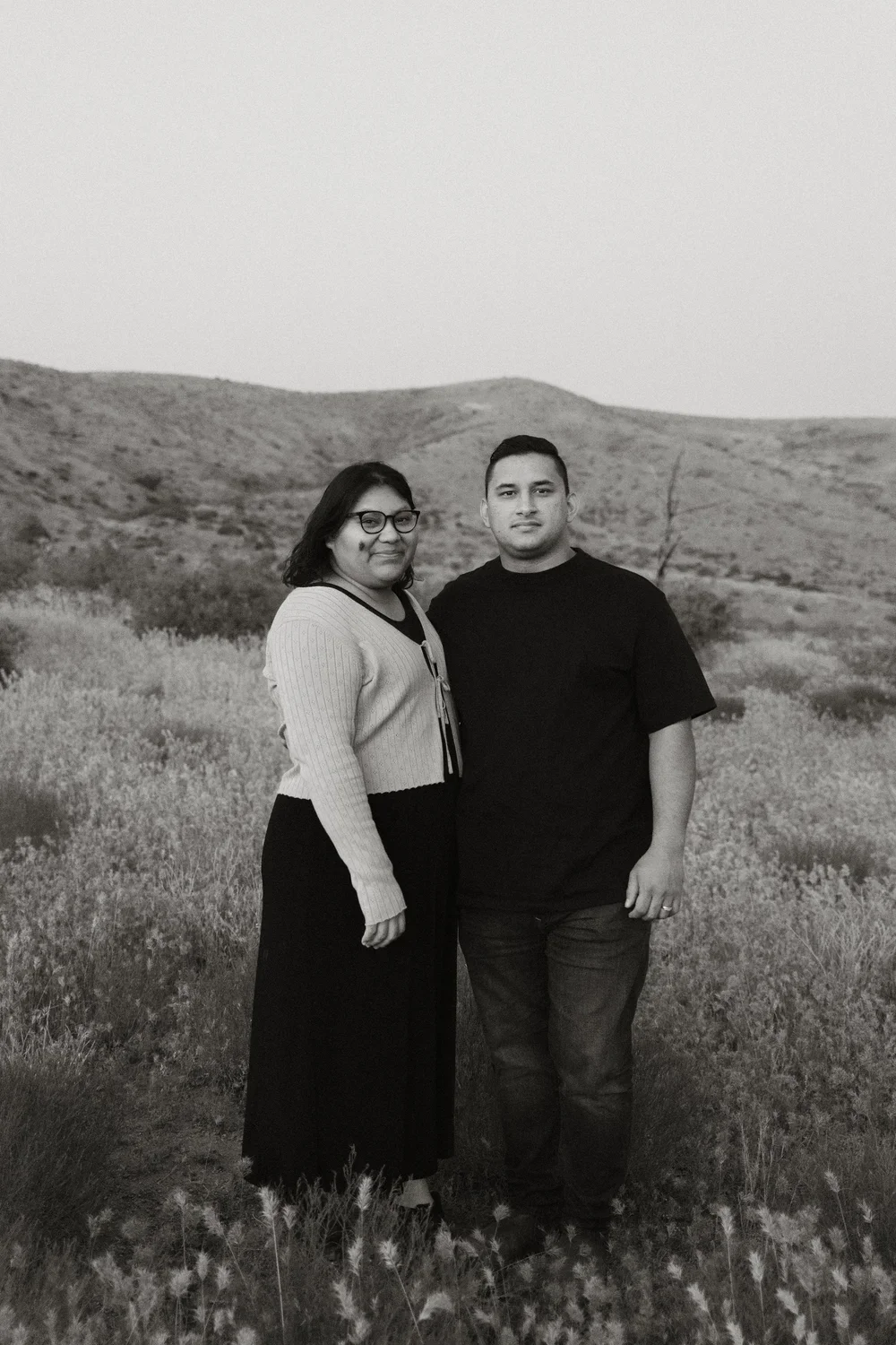 A black and white photo of two people standing outdoors in a field with hills in the background, a woman with glasses and a smile, and a man with short hair looking at the camera.