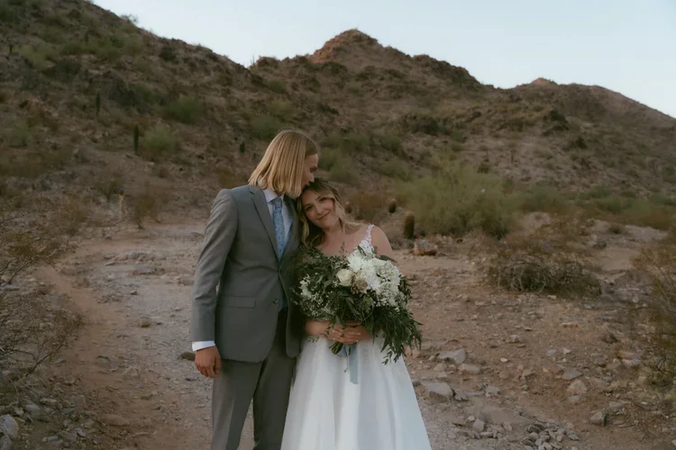 A bride and groom embrace outdoors in a desert landscape with hills in the background. The bride holds a large bouquet of white flowers and greenery. The groom is dressed in a gray suit and the bride in a white wedding dress.