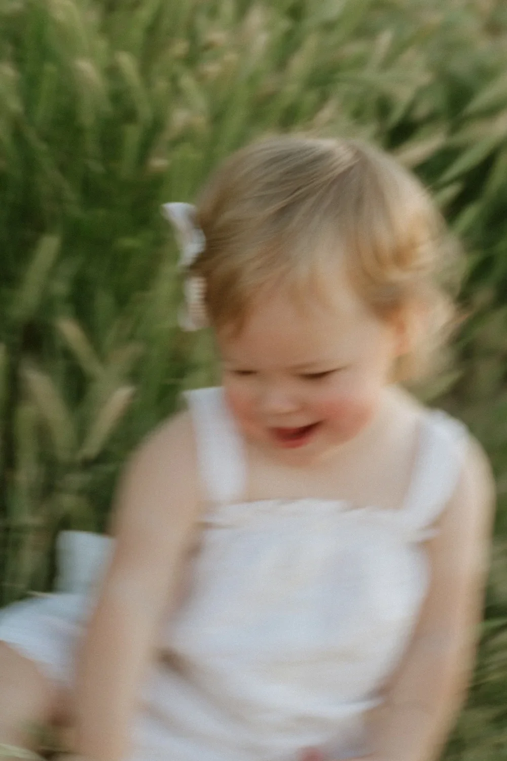 A young girl with blonde hair in a white dress smiling and playing outdoors amid tall green grass.