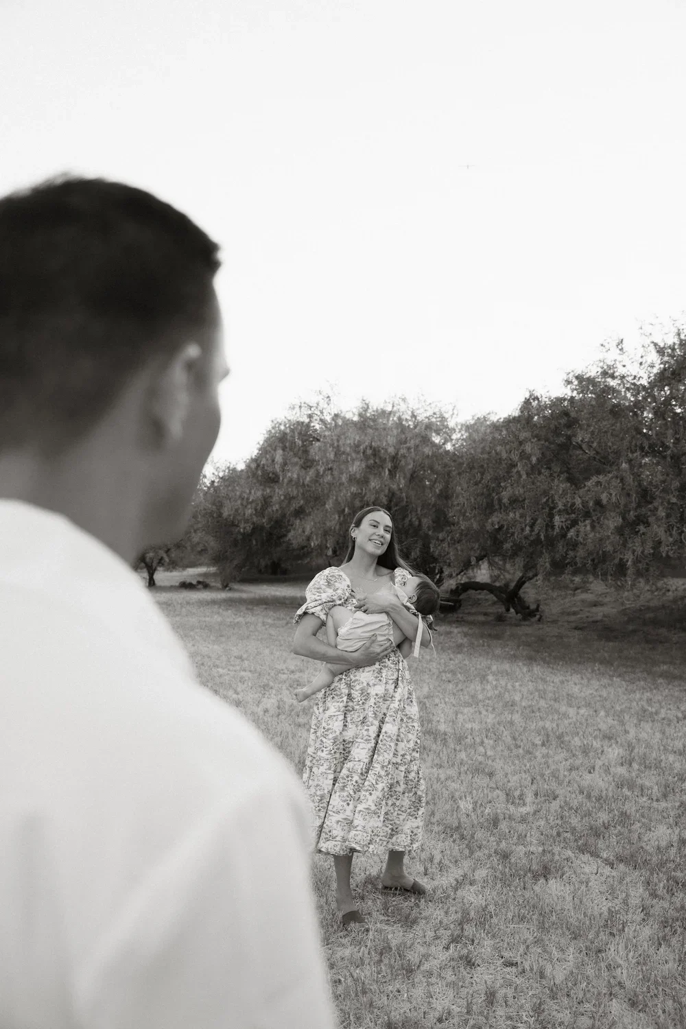 A woman smiling and holding a child outdoors with a man in the foreground, blurred, in a grassy area with trees.