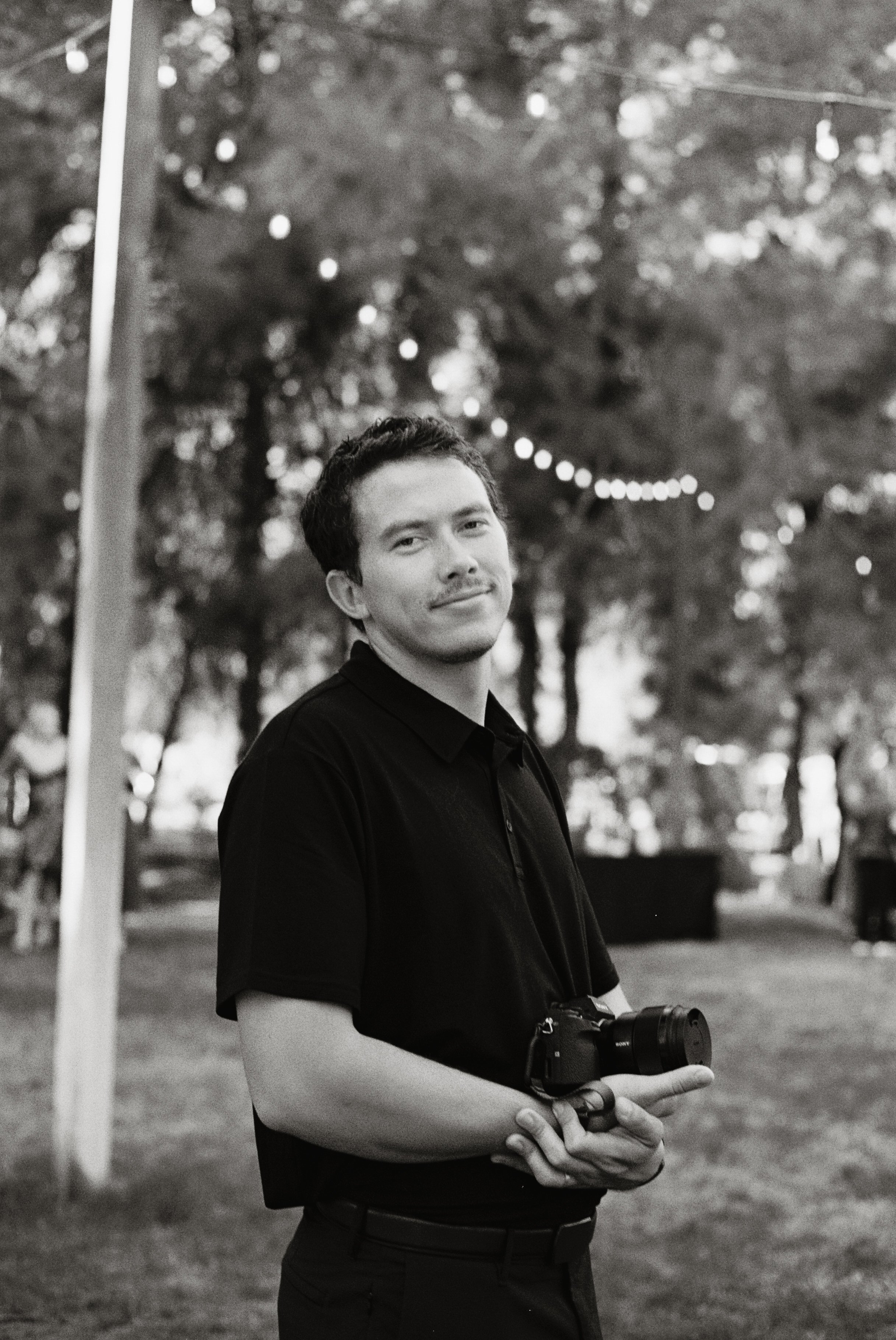 A young man with dark hair and a light mustache, wearing a dark shirt, stands outdoors in a park during the evening, holding a camera with both hands, with string lights and trees in the background.