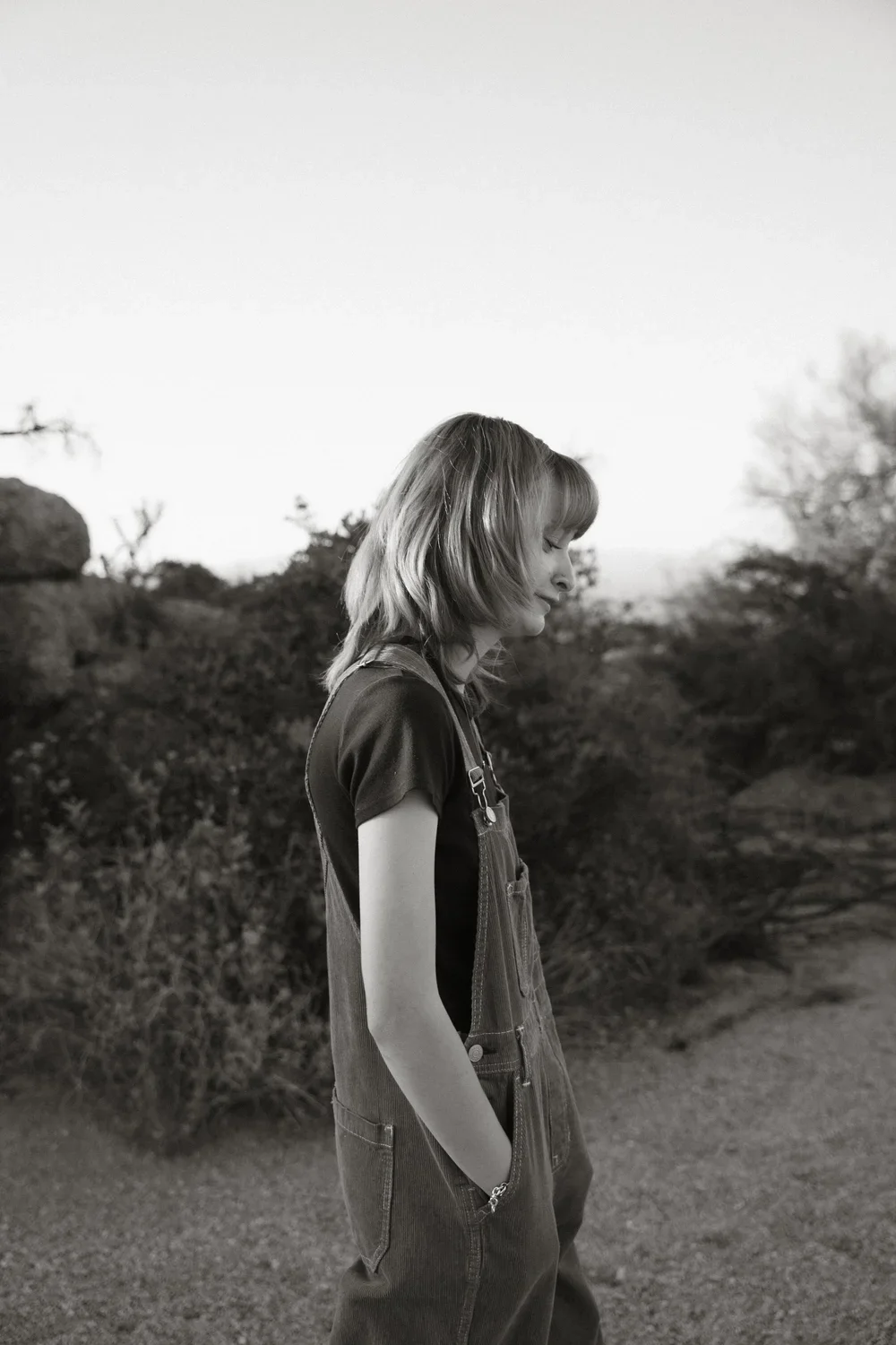 Black and white photo of a young woman standing outdoors, with her eyes closed and hands in her pockets, surrounded by trees and foliage.