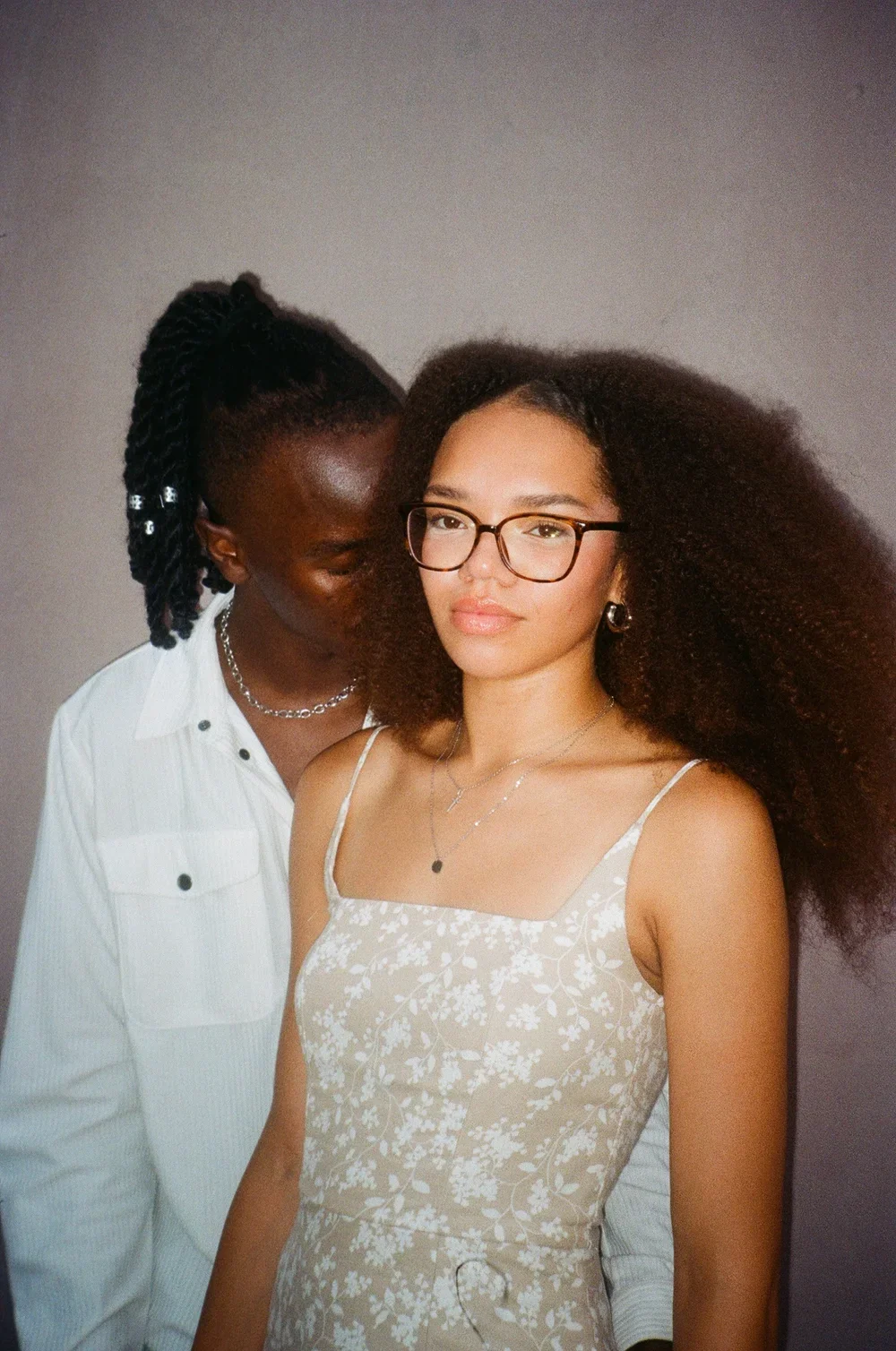 Two women, one with glasses and curly hair, and the other with dark skin and styled hair, standing close together against a gray background.