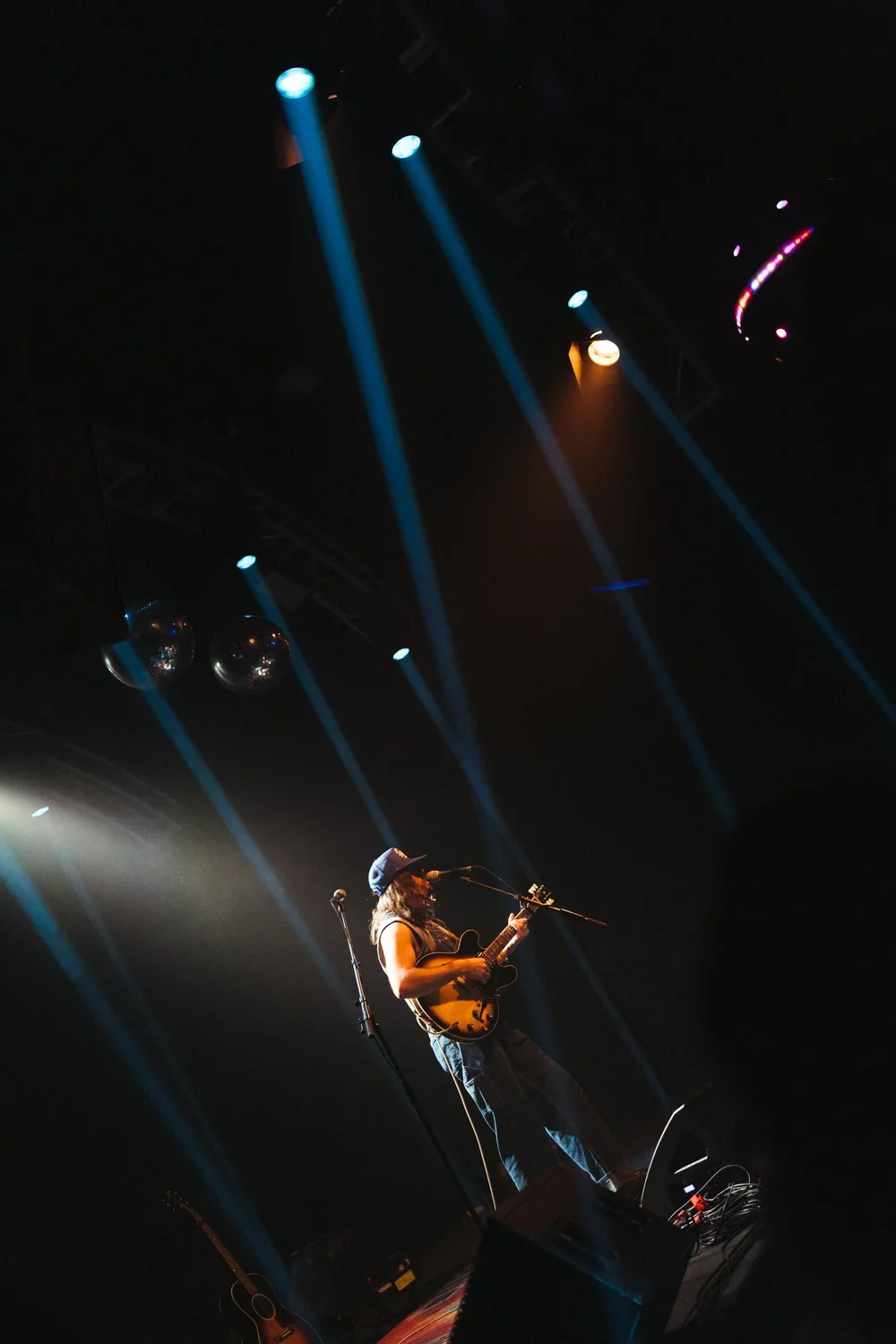 A musician playing an electric guitar and singing on stage under colorful spotlights at a concert.