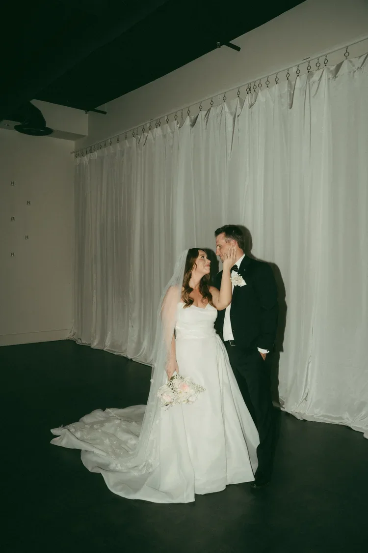 A bride and groom standing close together, gazing into each other's eyes, in front of a white curtain backdrop at their wedding.