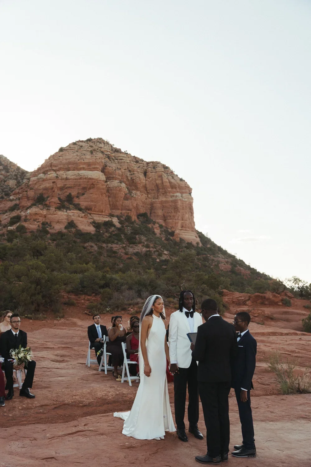 A wedding ceremony taking place outdoors in a desert with red rock formations background. The bride is wearing a white gown and veil, and the groom is in a white tuxedo jacket. Two men are officiating the ceremony, and guests are seated behind them.