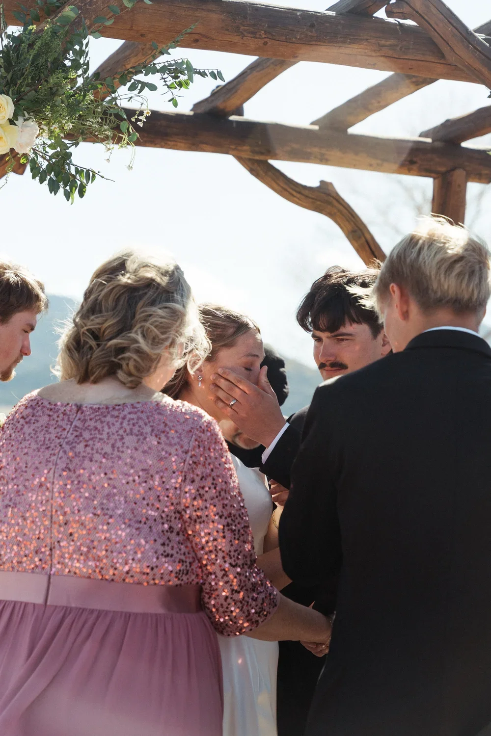 A wedding ceremony outdoors with a wooden arch decorated with flowers and greenery. A woman, possibly the bride, is emotional while a man, possibly the groom, touches her face. Other individuals surround them, witnessing the moment.
