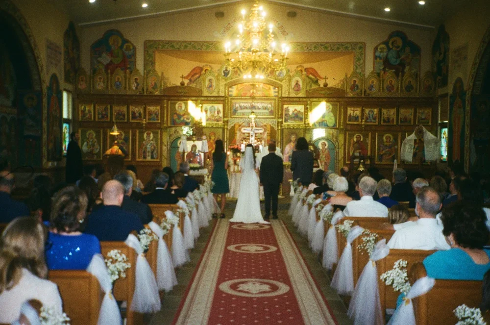 A wedding ceremony inside a church with a congregation seated on both sides, the altar decorated with religious icons, and a chandelier hanging from the ceiling.