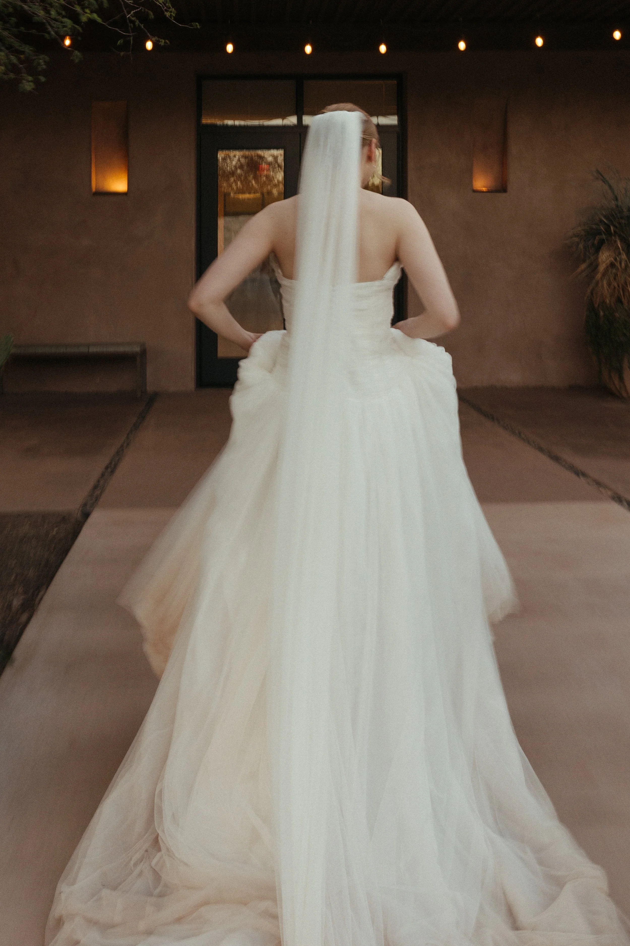 Woman in a white wedding dress with a long veil standing outside at night, facing away from the camera, with string lights overhead and a building with a glass door in the background.