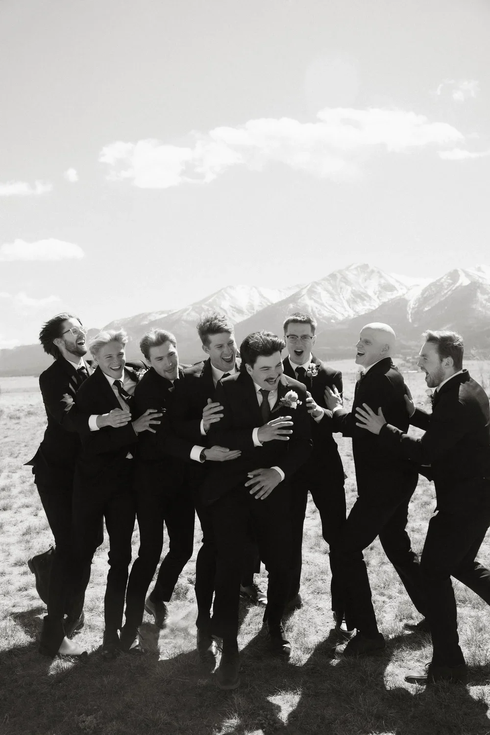 A group of men in suits laughing and celebrating outdoors with mountains in the background.