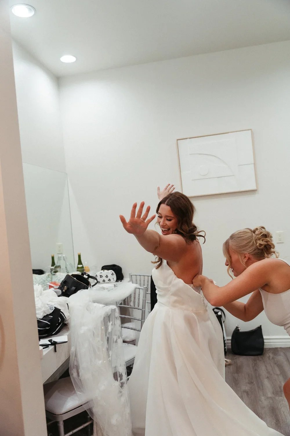 A woman in a white wedding dress smiling and playfully pushing away another woman helping her with her dress, in a room with white walls and a table cluttered with various items.