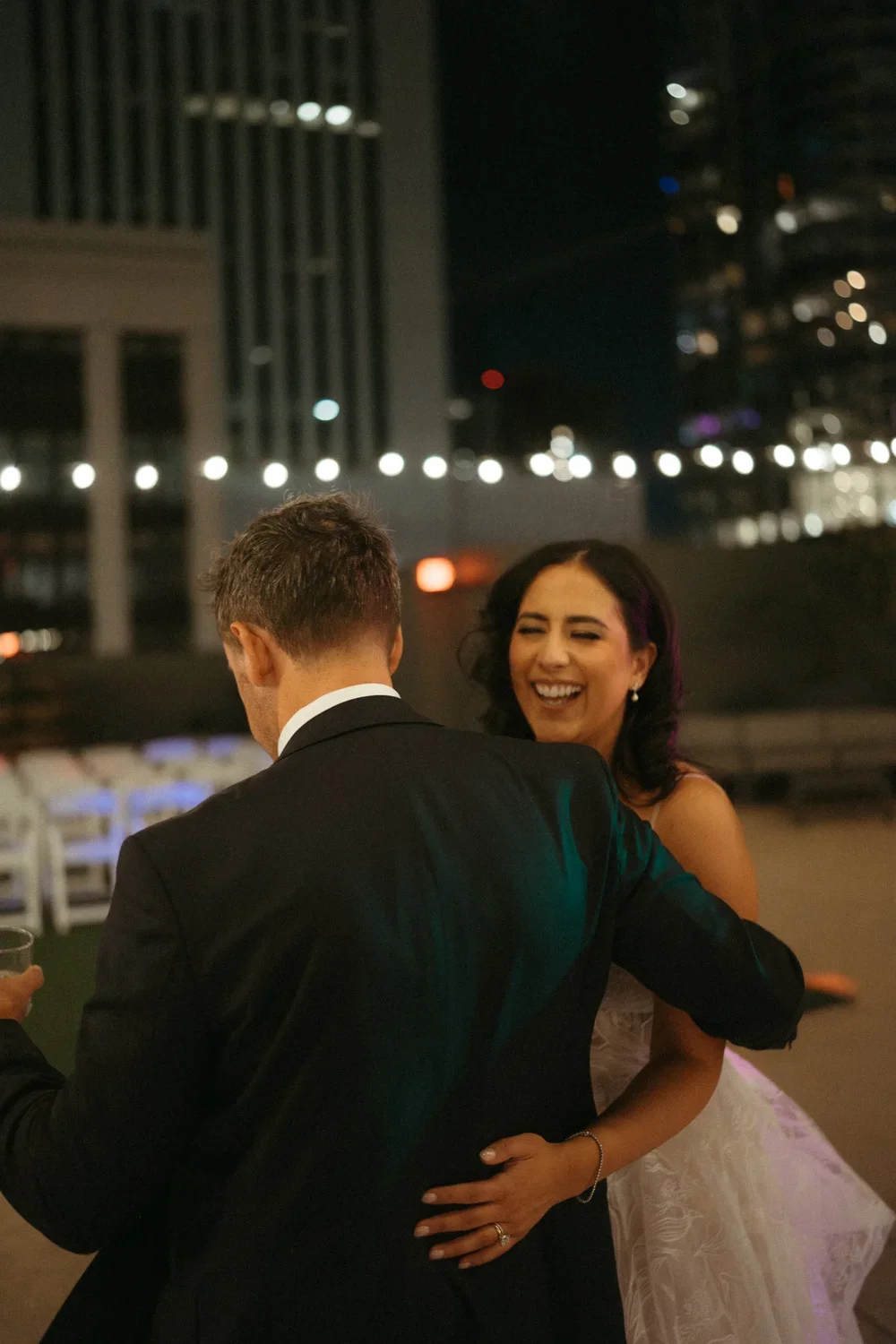 A woman in a white wedding dress and a man in a black suit dancing and smiling at a nighttime outdoor wedding reception with city lights in the background.
