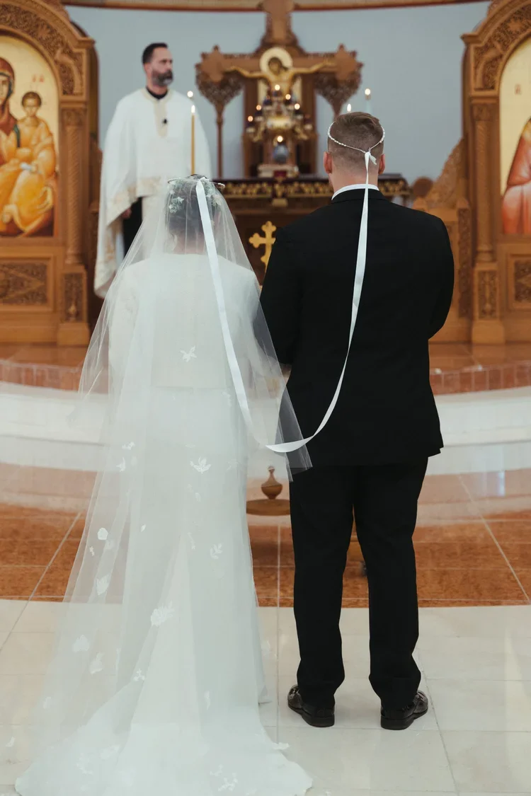 A bride and groom standing at the altar during a wedding ceremony in a church, with the priest officiating, religious icons on the walls, and the bride wearing a veil and wedding gown, while the groom is in a black suit.