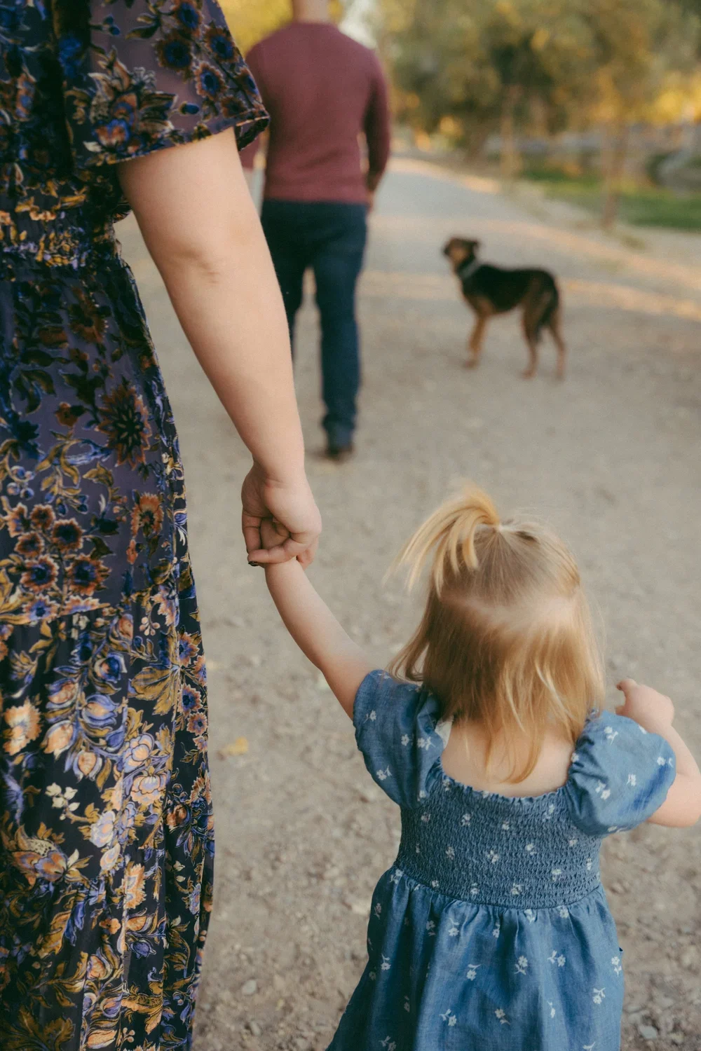 A woman holding a young girl’s hand while walking outdoors on a dirt path, with two dogs in the background.