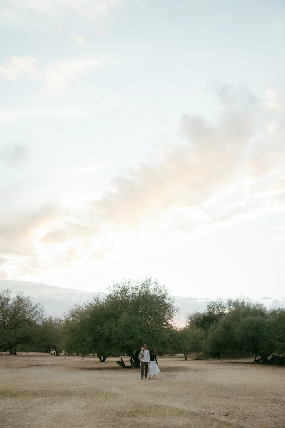 A couple walking hand in hand in a dry, open field with sparse trees during sunset in a rural area.