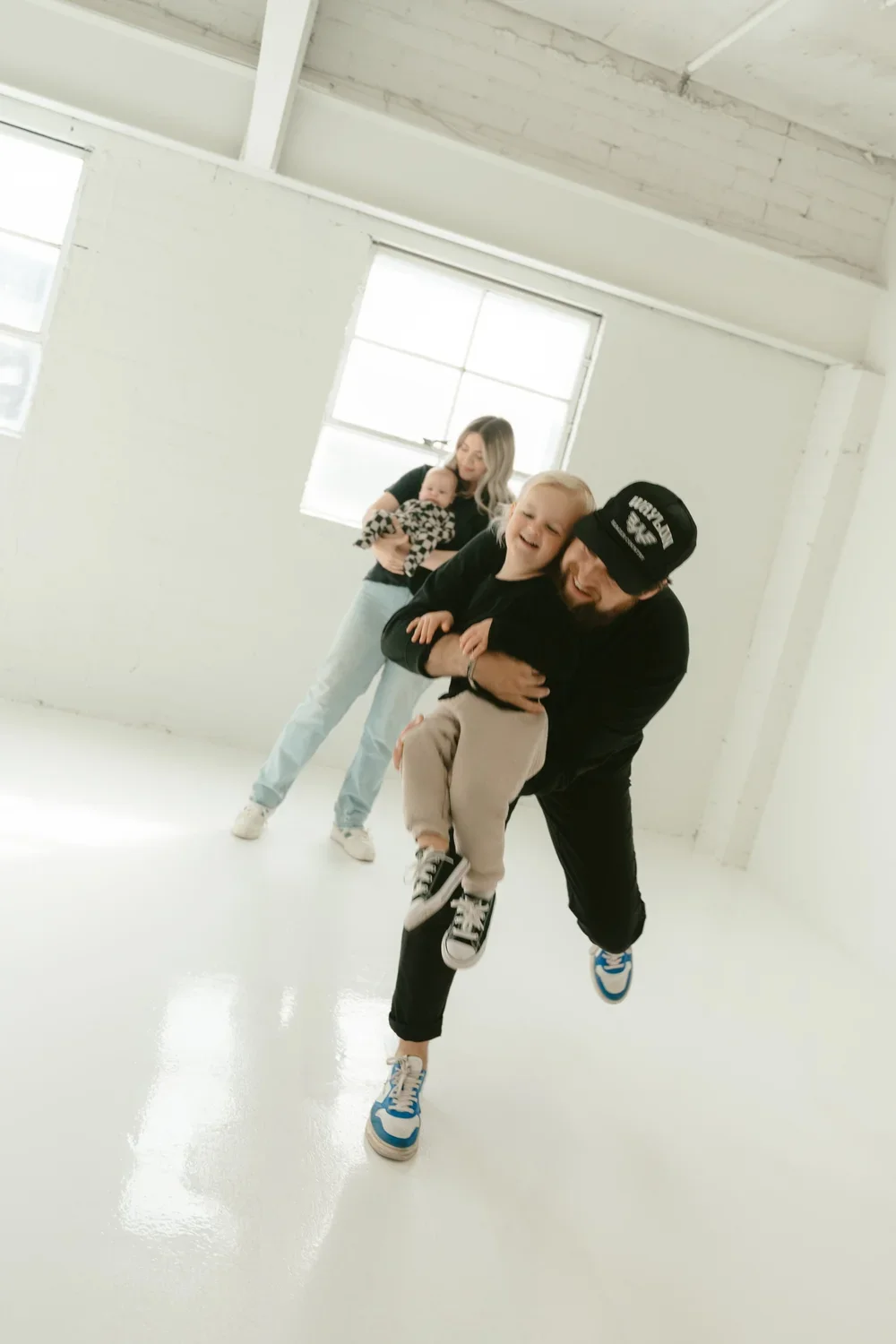 A man is playfully lifting a young boy in an empty, white-walled room with large windows, while a woman stands in the background holding a baby and watching them.