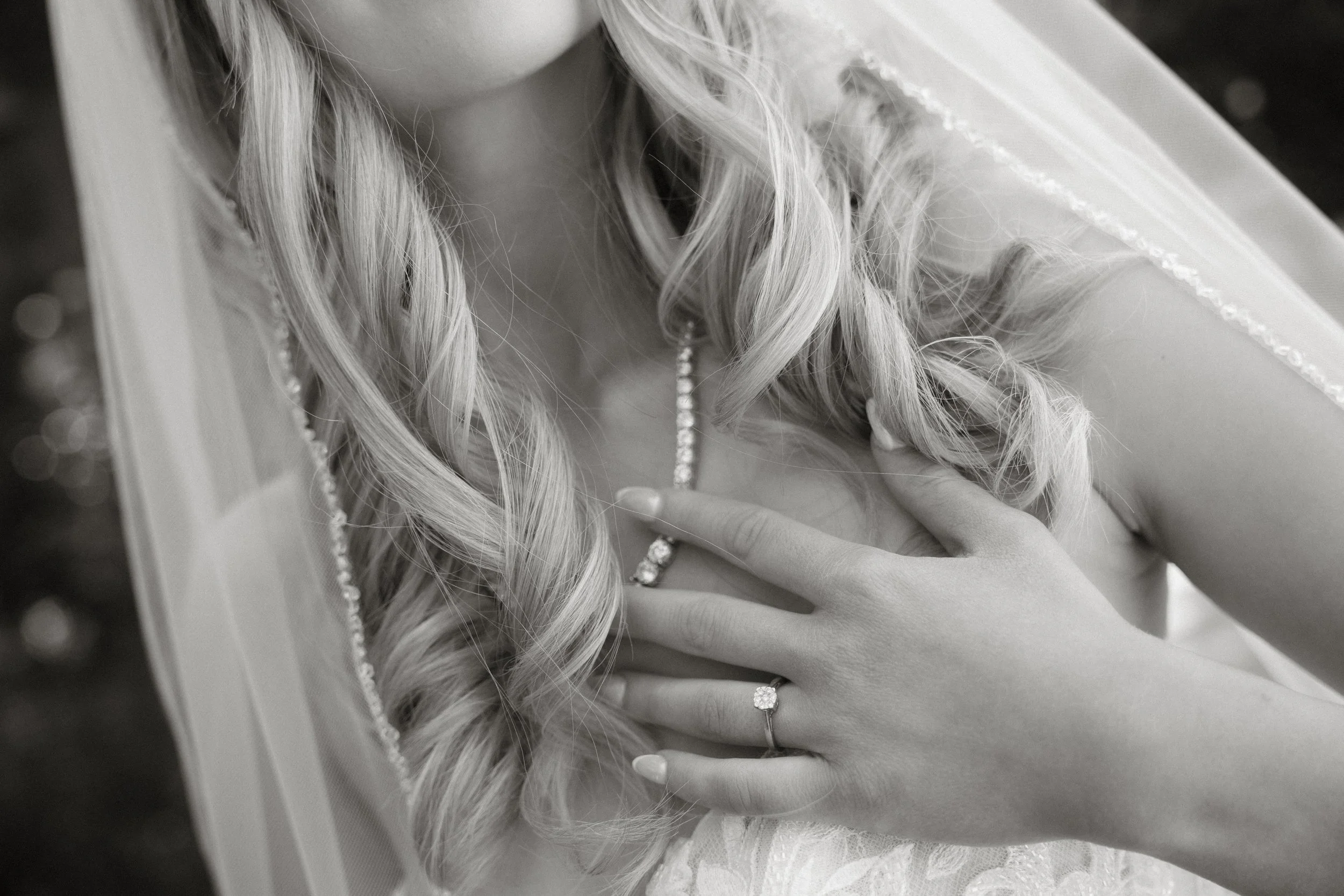 A close-up black-and-white photo of a woman with long, wavy blonde hair, wearing a diamond ring, necklace, and dress, touching her chest with her hand.