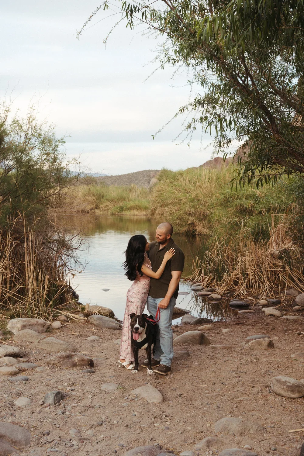A couple and their dog standing on rocky ground near a river with bushes and trees around, with a mountain in the background.