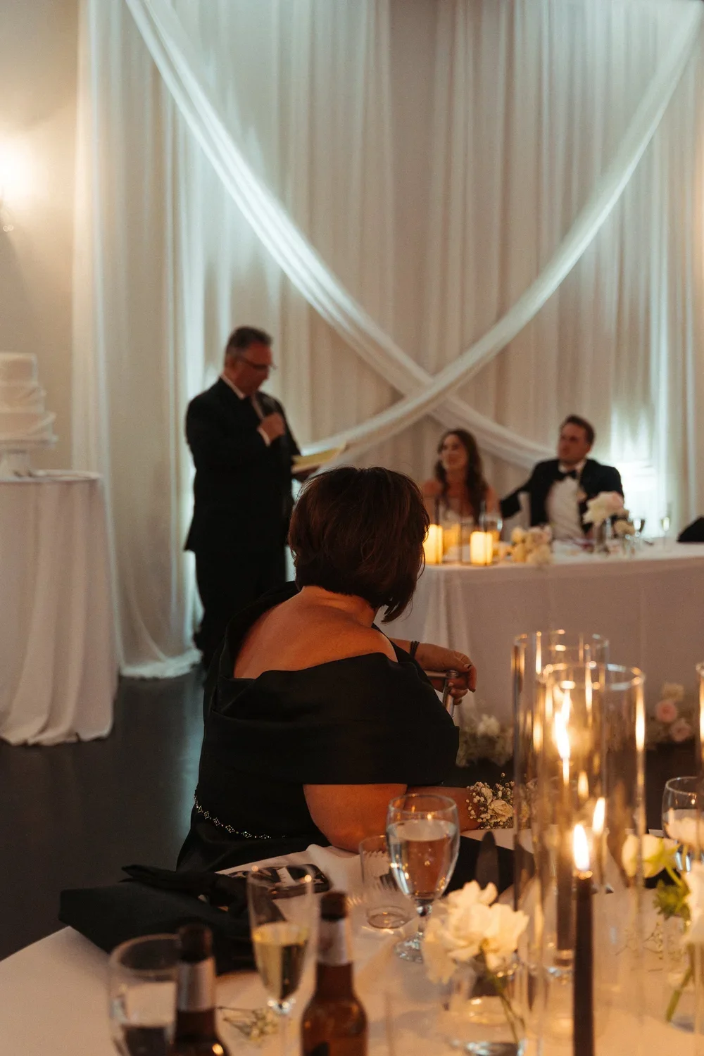 A wedding reception with a seated woman in the foreground, a man giving a speech in the background, and a bride and groom sitting at a decorated head table with candles and floral arrangements.